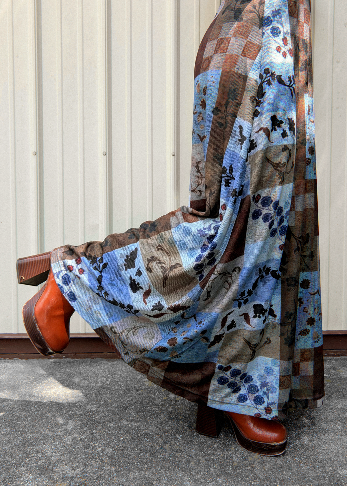 A person wearing a long patchwork print dress in shades of blue, brown, and beige, paired with brown leather boots, standing against a corrugated metal wall.