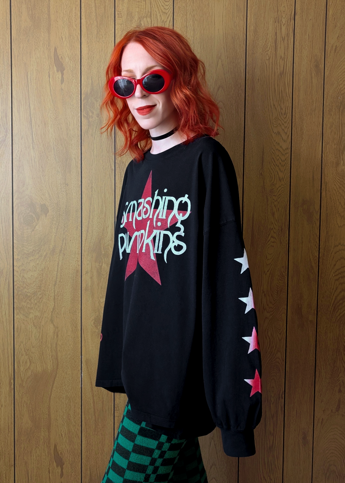 Person wearing a black long sleeve shirt with Smashing Pumpkins star designs, standing against a wooden panel background.