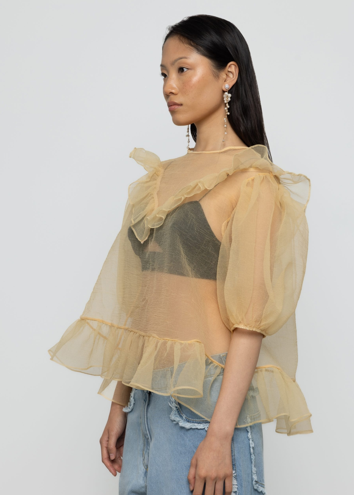 Woman wearing a sheer yellow organza balloon sleeve top over a dark bralette with denim jeans on a white background