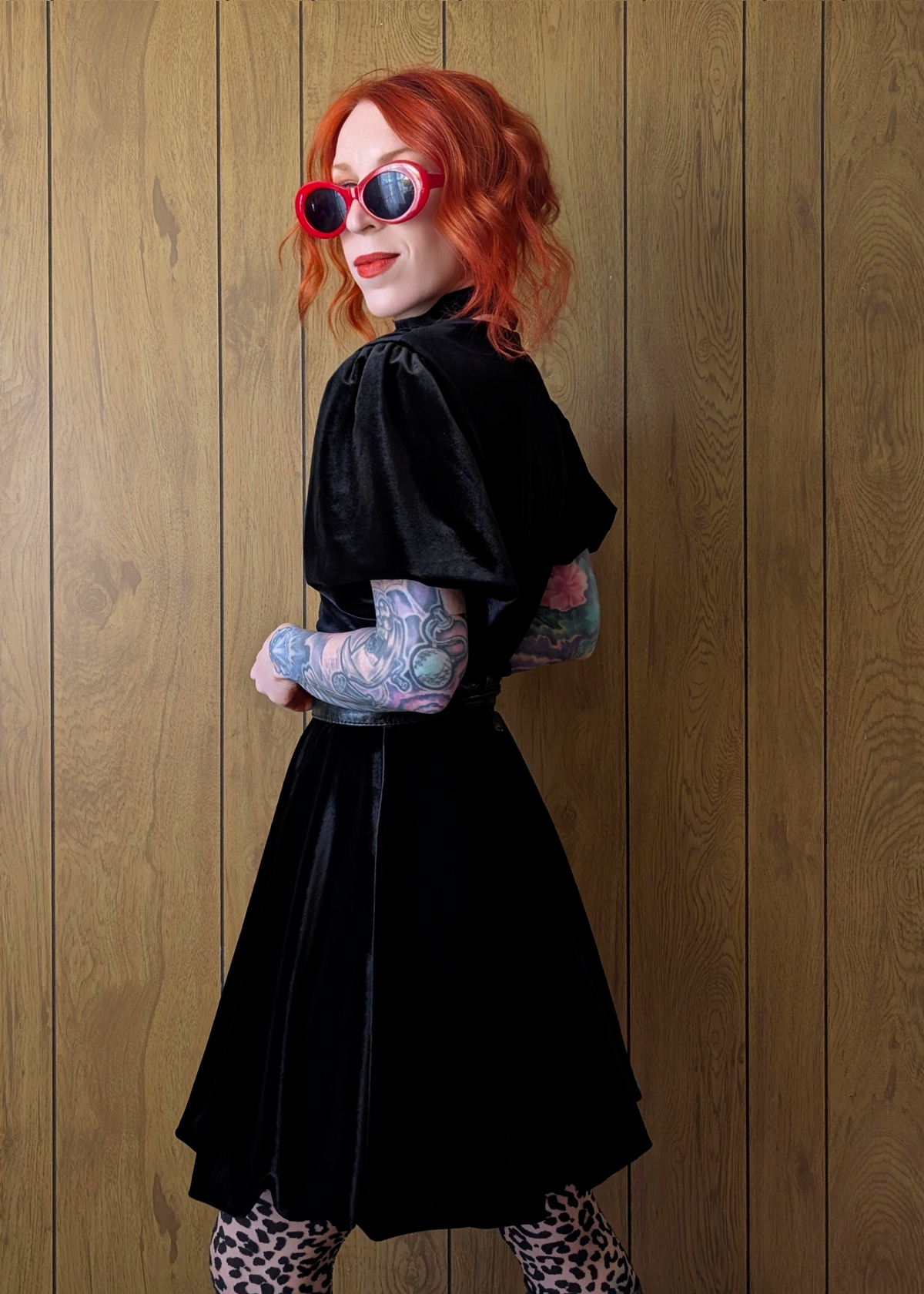 Person with red hair wearing a black velvet short sleeve dress and sunglasses against a wooden panel background