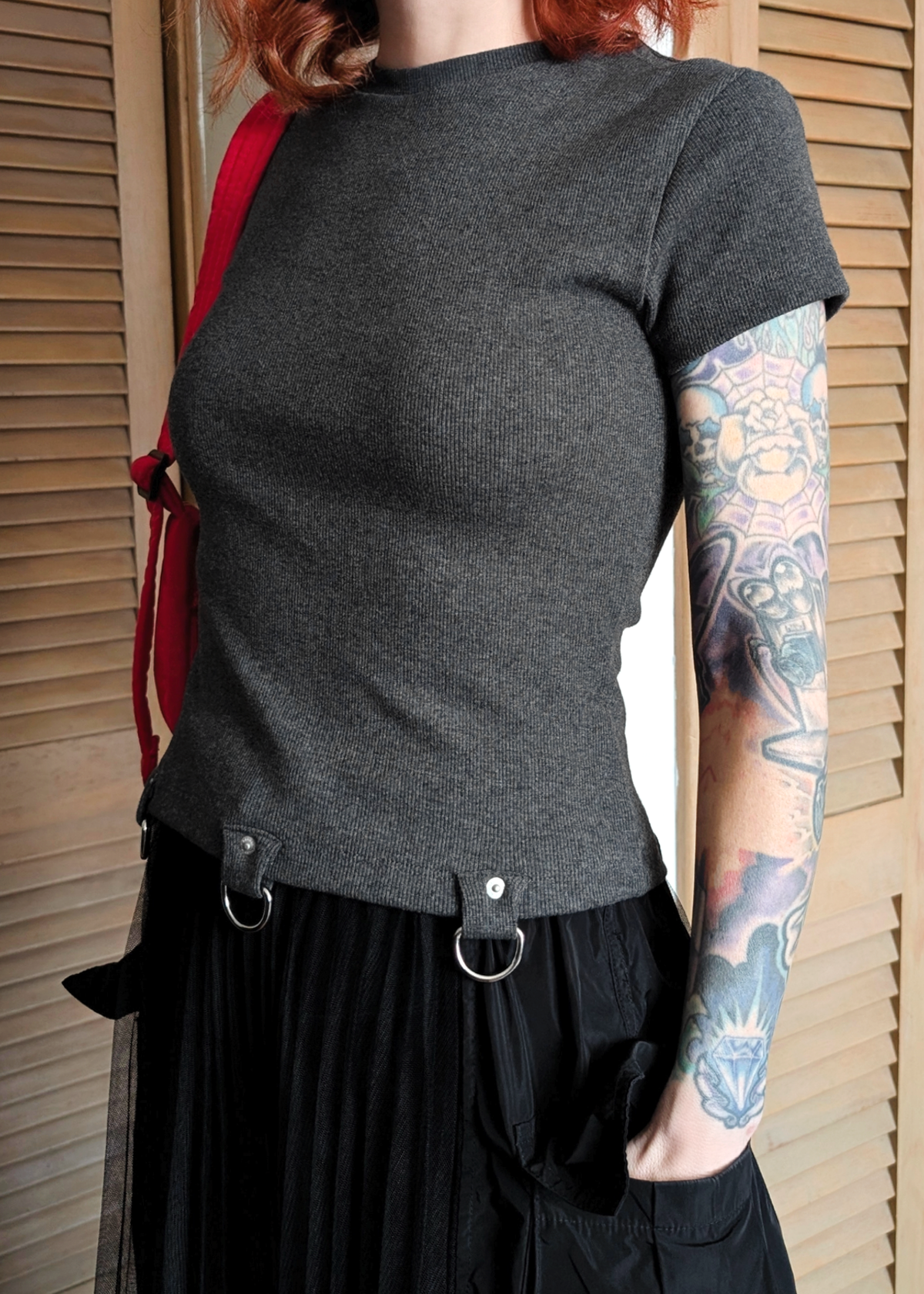 Woman wearing a ribbed charcoal grey t-shirt with D-ring details at hem, and black skirt standing indoors.