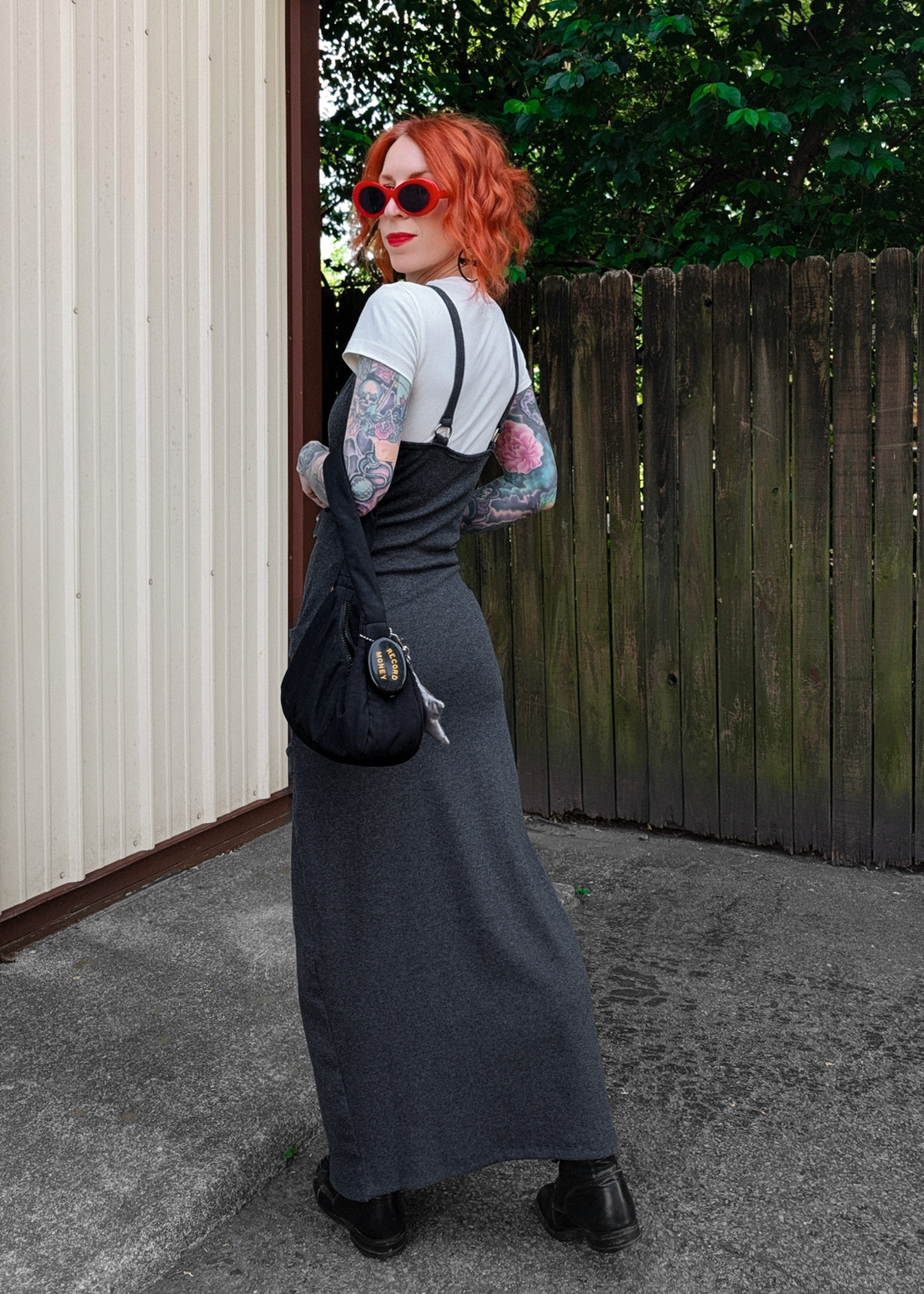 Person with red hair wearing a midi-length charcoal rib dress and white tee and carrying a black bag, standing in front of a wooden fence.