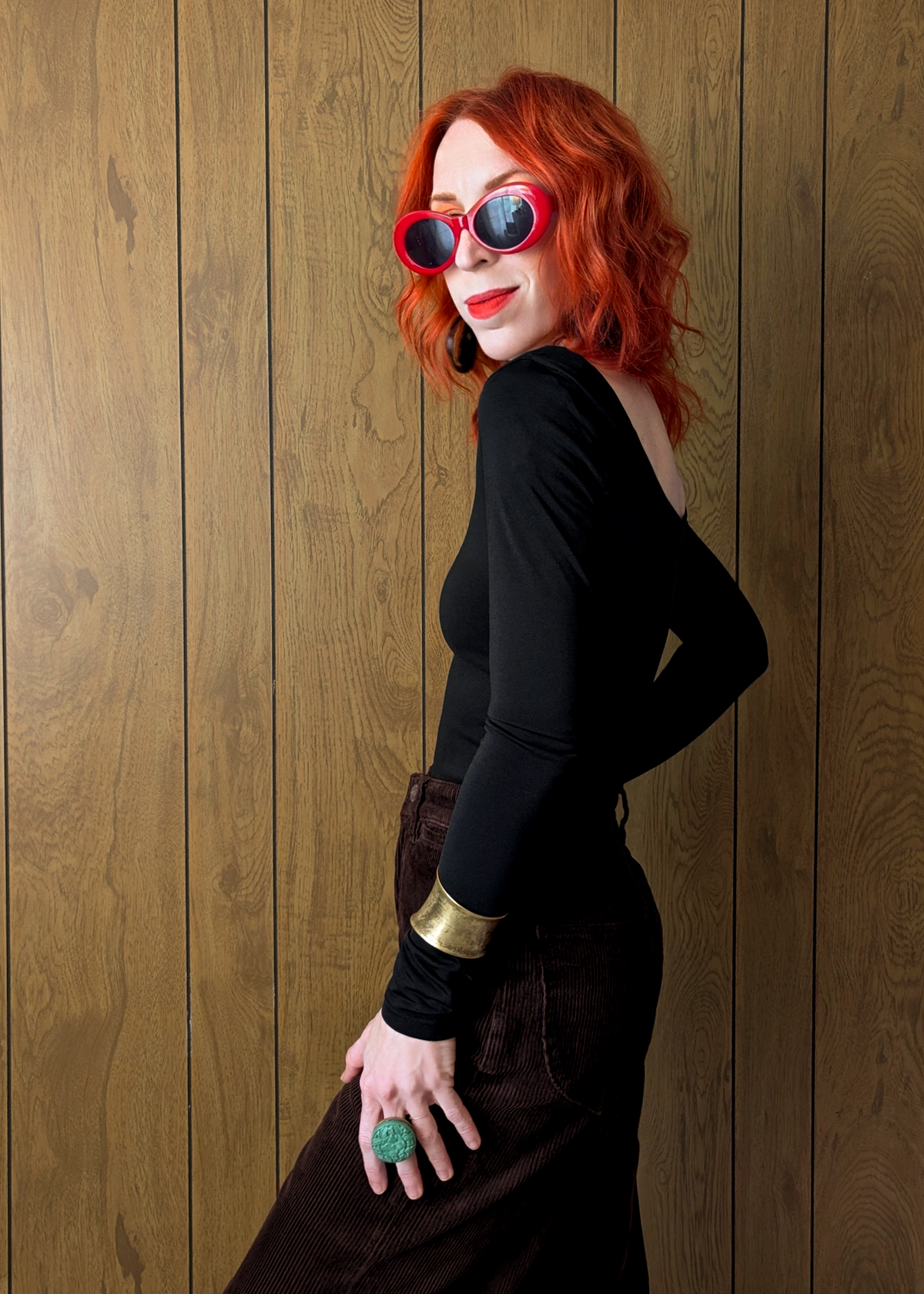 Person with red hair wearing sunglasses and a black top and brown cords against a wooden panel background
