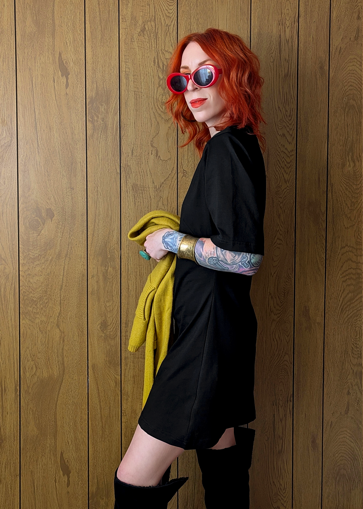 Person with red hair wearing sunglasses and a black mini t-shirt dress against a wooden wall 
