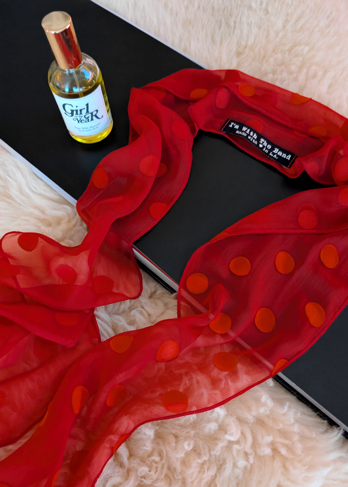 Lifestyle photo of a red polka dot scarf laid flat on a cream faux fur rug with a black book and bottle of perfume