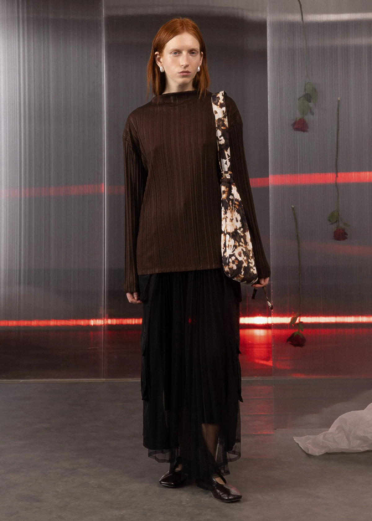 Model wearing a brown top and black sheer mesh cargo skirt with a patterned bag in a minimalistic setting.