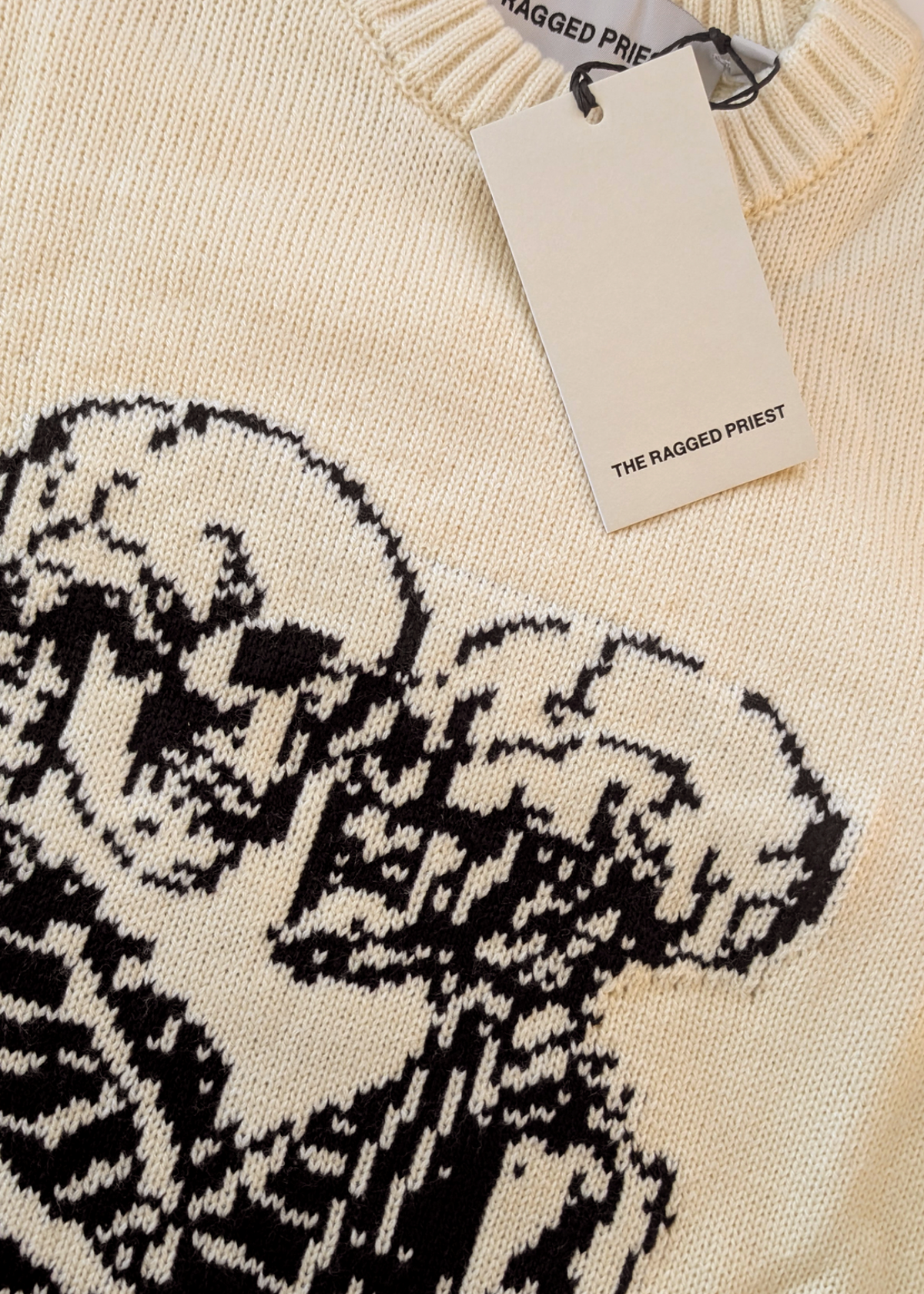 Knitted sweater with black design and 'The Ragged Priest' tag on beige background