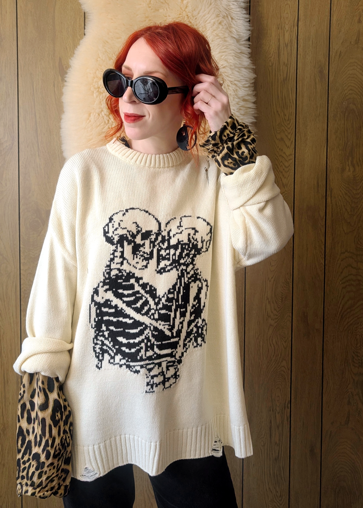 Person wearing a cream sweater with a black skeleton design, holding sunglasses, against a wooden wall.