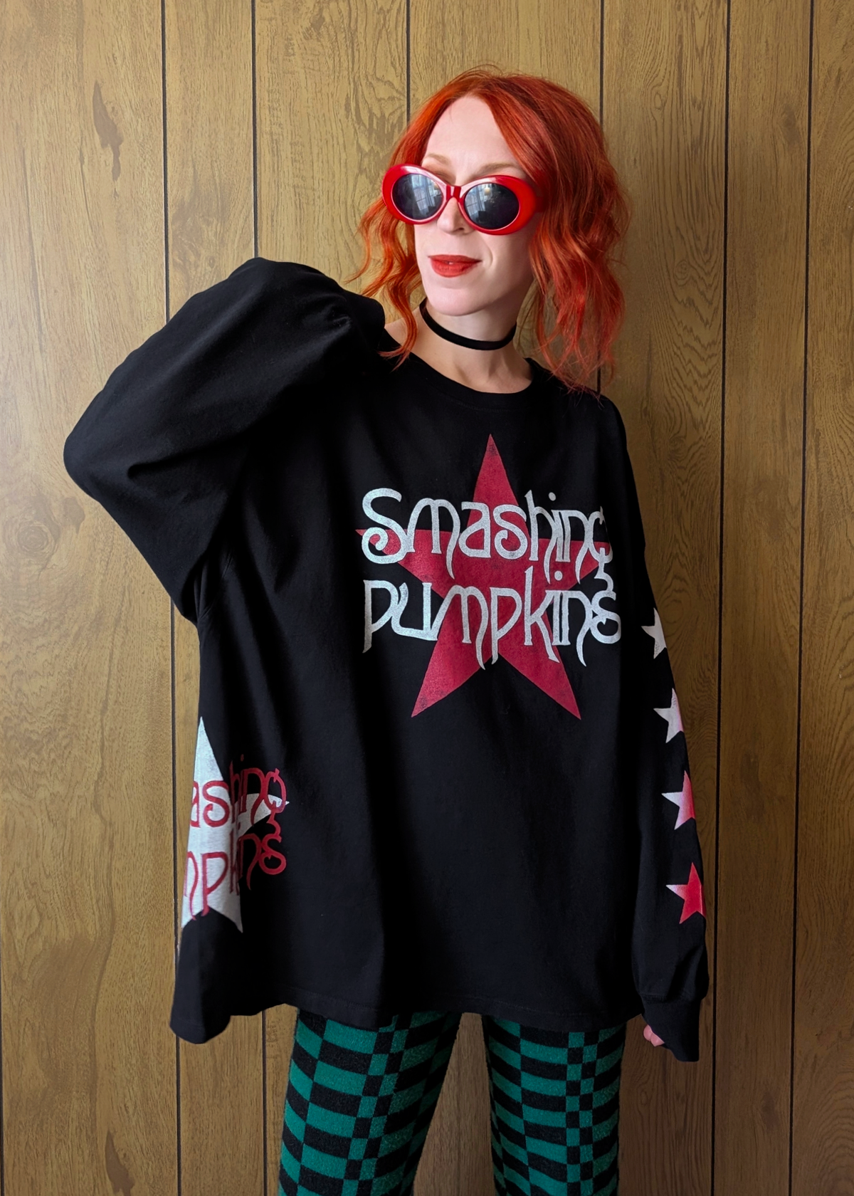 Person wearing a black 'Smashing Pumpkins' oversized long sleeve shirt with red star design against a wooden wall.