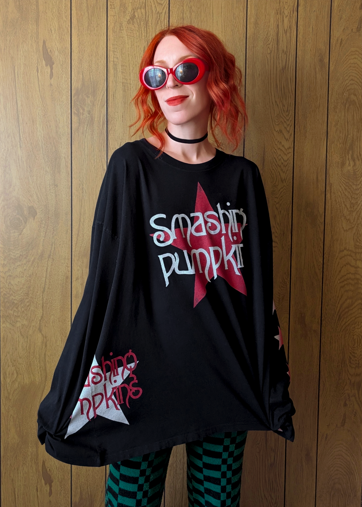 Person wearing a black oversized t-shirt with 'Smashing Pumpkins' logo, red sunglasses, and striped pants against a wooden panel background.