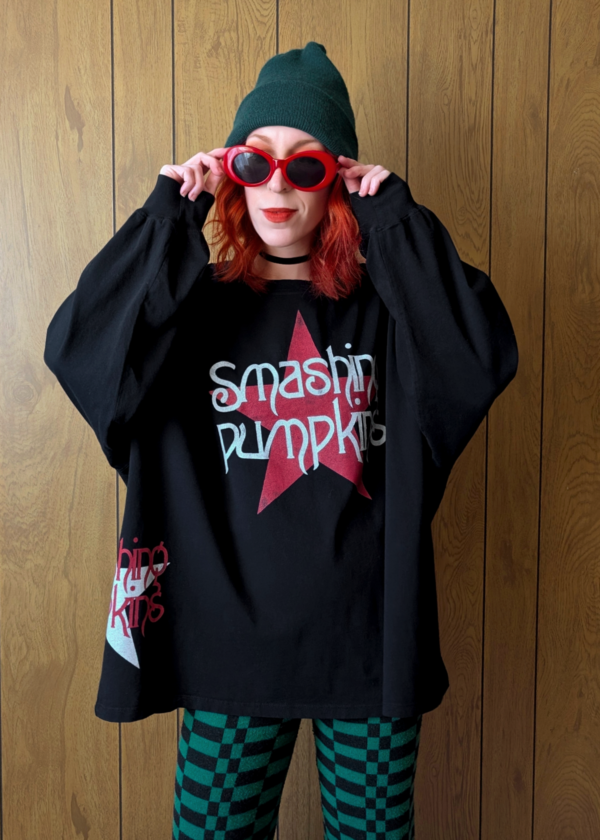 Person wearing a black oversized long sleeve shirt with 'Smashing Pumpkins' logo, red sunglasses, and checkered pants against a wooden panel background.