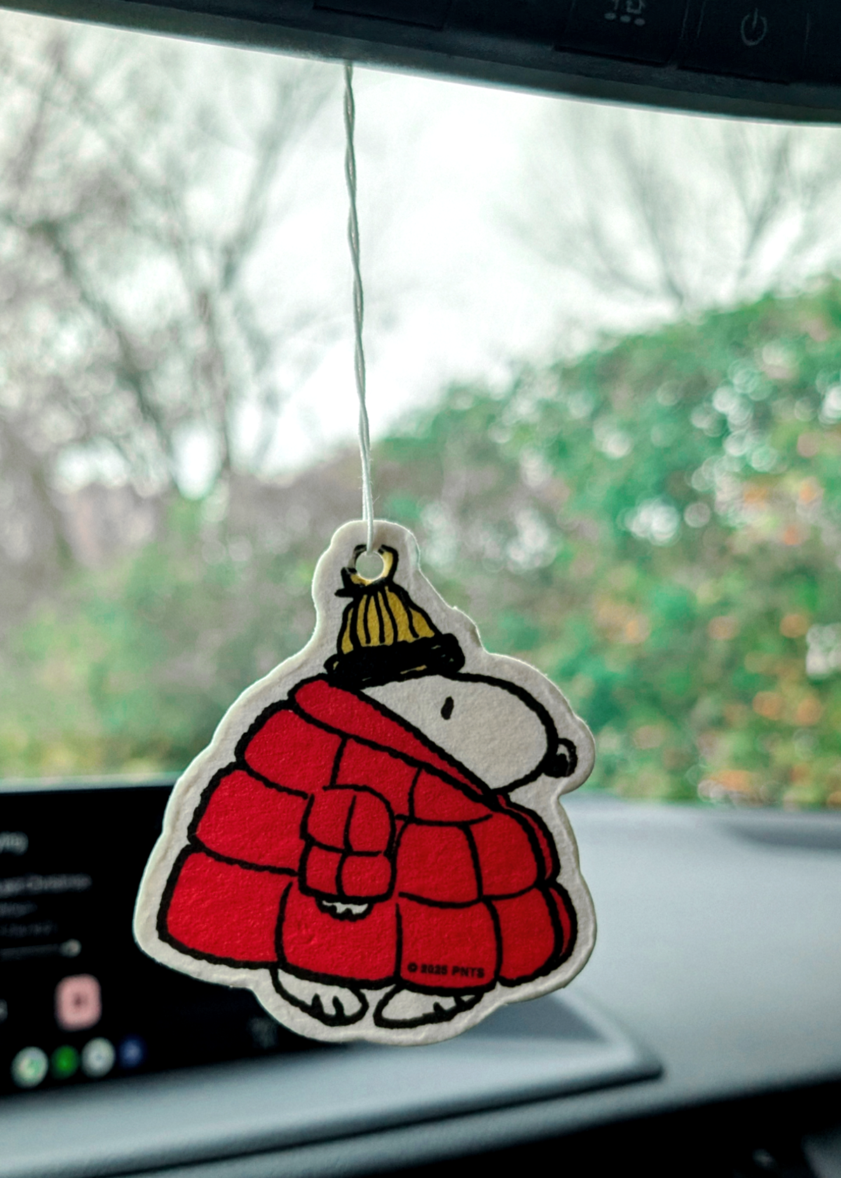 Car air freshener shaped like a Snoopy in a red coat and yellow hat hanging from a car's rearview mirror.