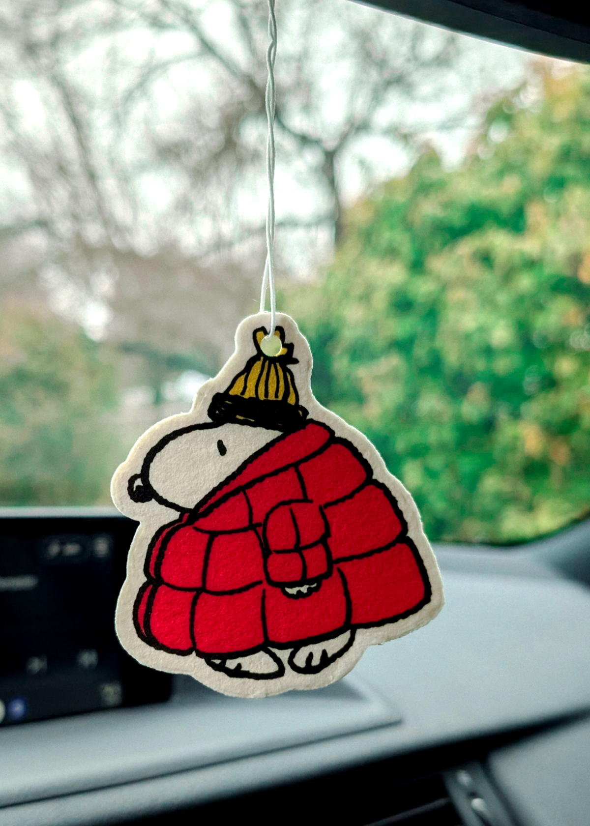 Car air freshener shaped like a Snoopy in a red coat and yellow hat hanging from a car's rearview mirror.