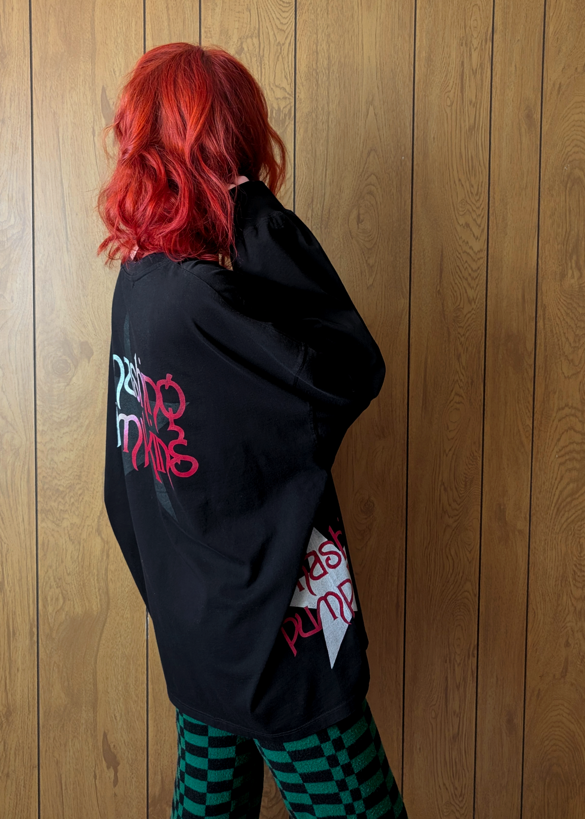 Person wearing a black long sleeve t-shirt with red and grey Smashing Pumpkins design against a wooden wall