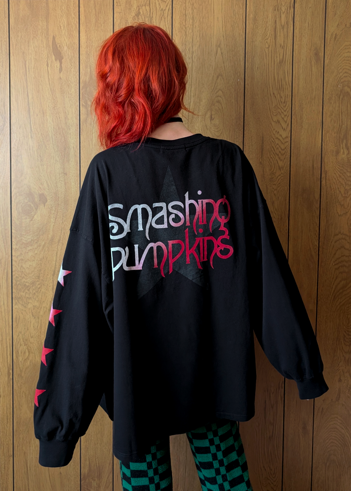 Person wearing a black oversized sweatshirt with 'Smashing Pumpkins' text on the back, standing against a wooden panel wall.