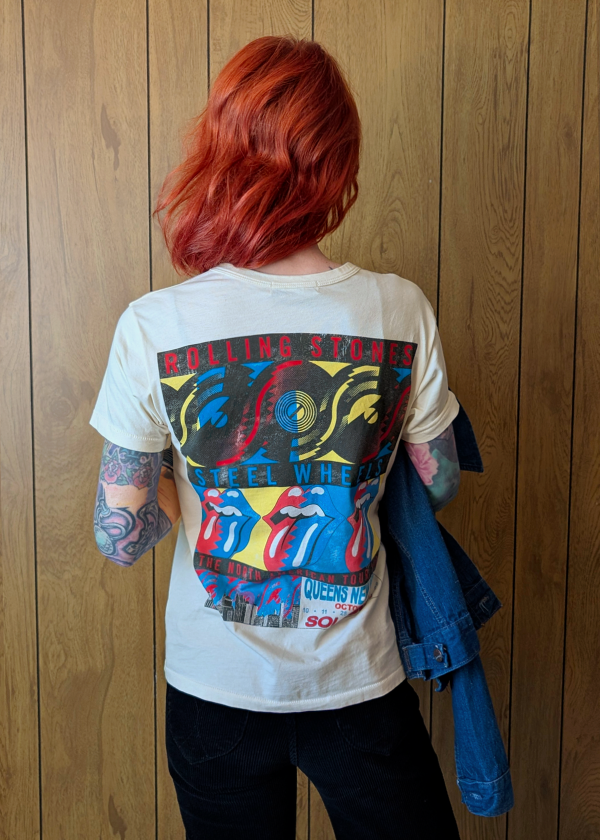 Person wearing a cream Rolling Stones t-shirt with colorful graphics on the back, standing against a wooden wall.
