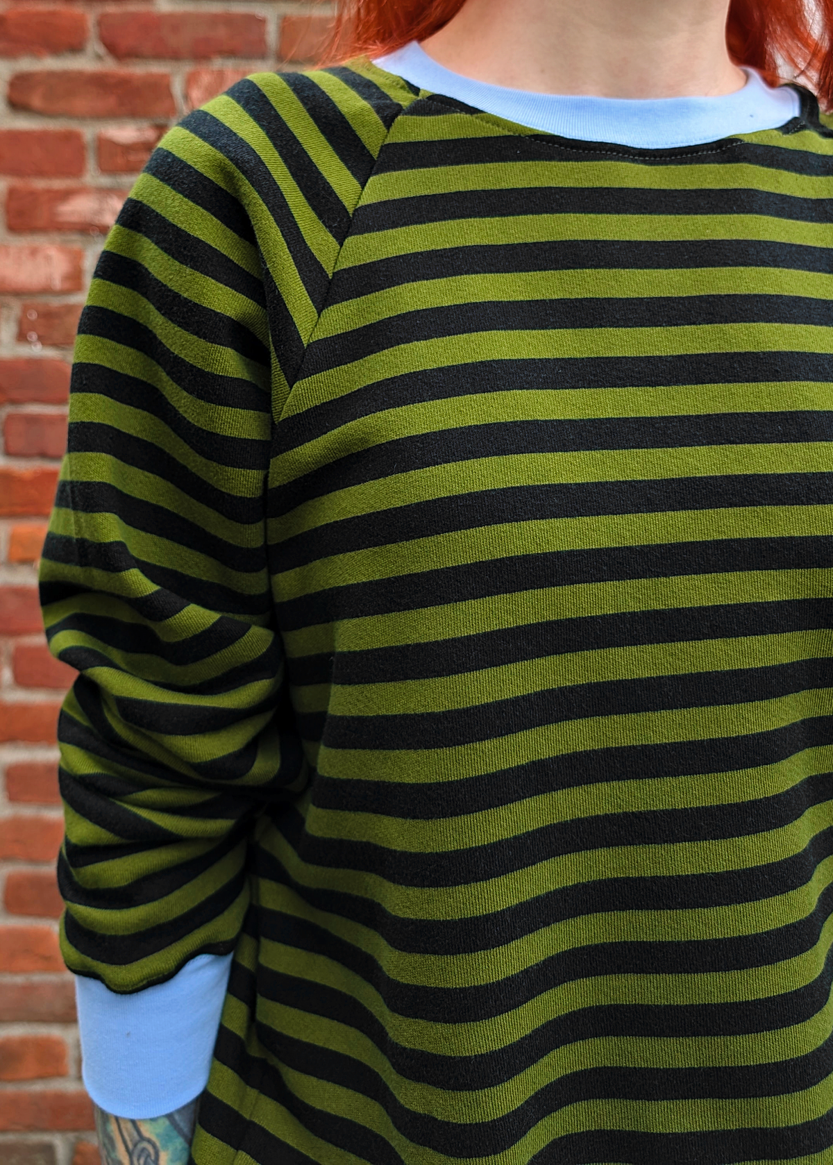 Details of the Olive green and black stripe raglan crew neck sweatshirt with ribbed sky blue cuffs, collar, and hem. Slouchy, relaxed fit. Made of 100% french Terry cotton