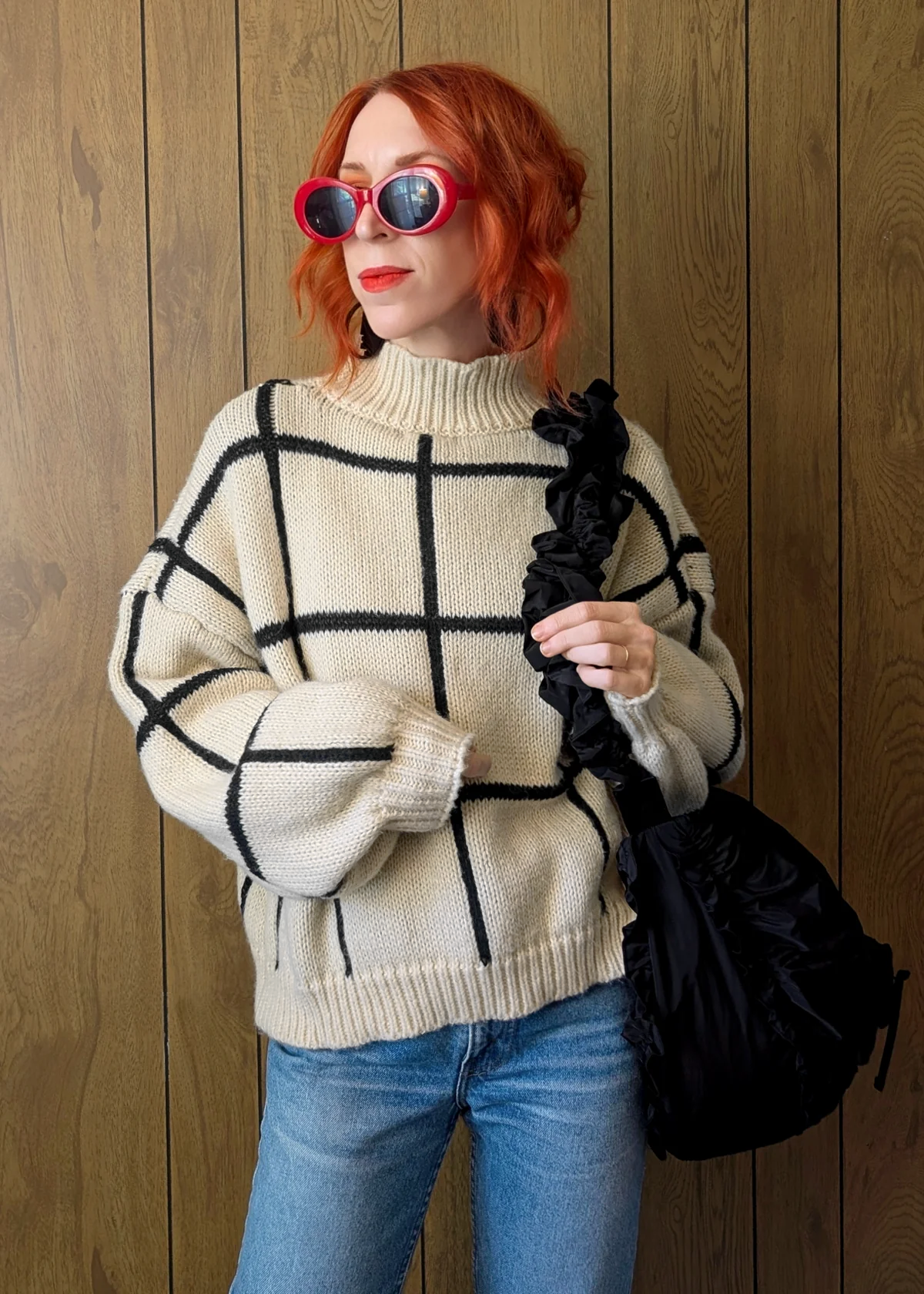 Person wearing a checkered sweater and sunglasses, holding a black bag against a wooden background
