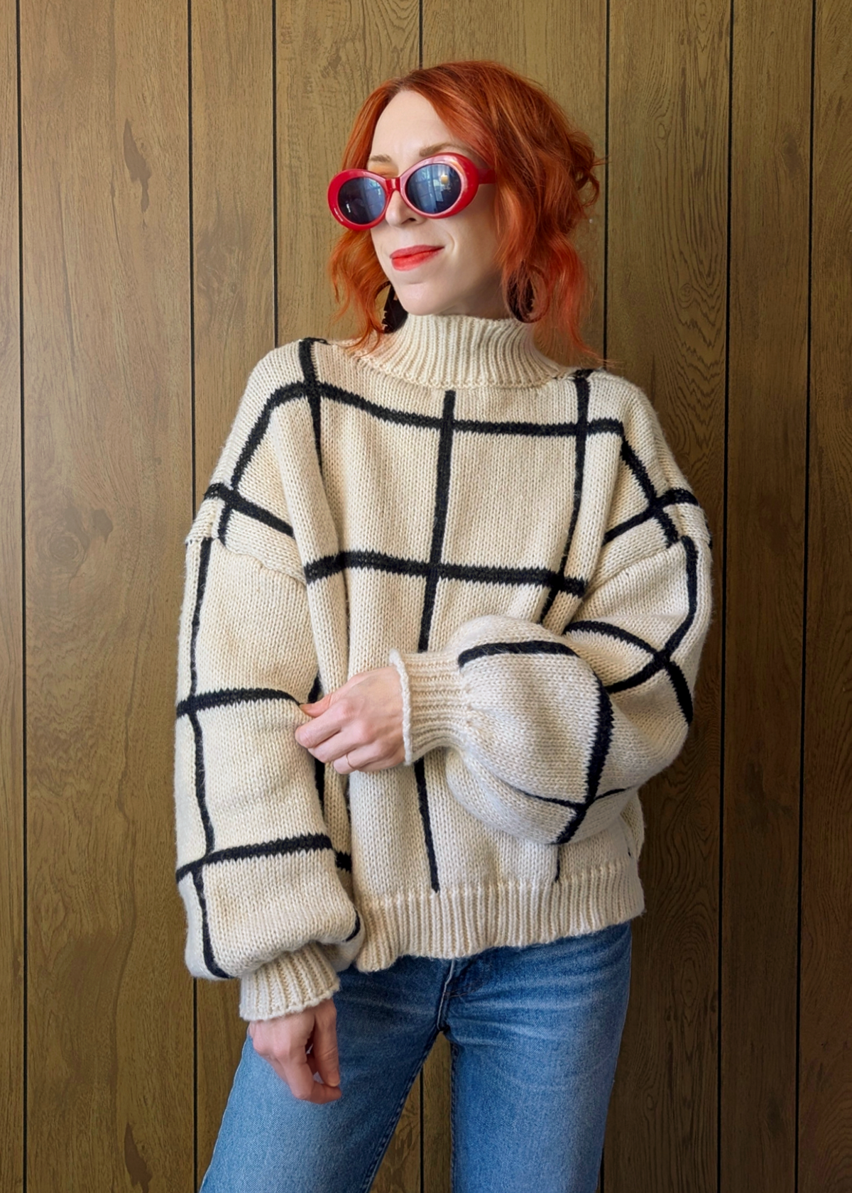 Person wearing a cream sweater with black grid pattern and blue jeans against a wooden background
