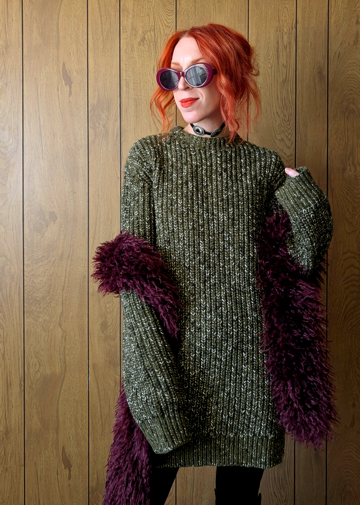 Person wearing a green knitted sweater styled with a fuzzy purple scarf against a wooden background