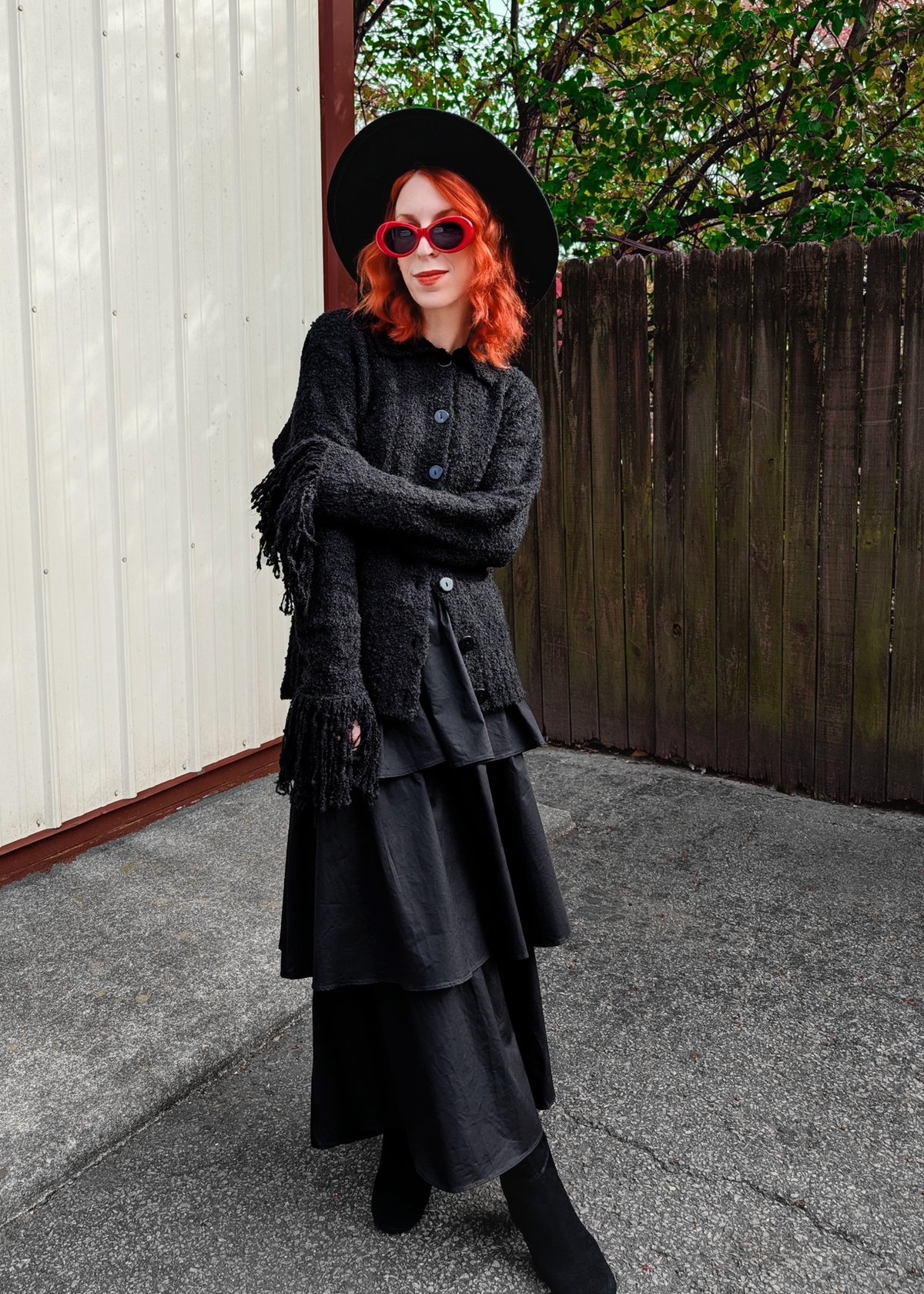 Person wearing a black cardigan, hat, and tiered ruffle skirt with red hair and sunglasses outdoors.