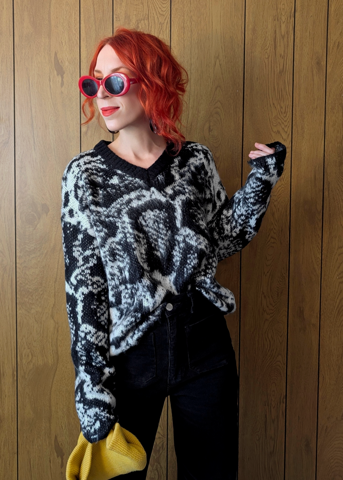 Person with red hair wearing a patterned sweater and sunglasses against a wooden wall.