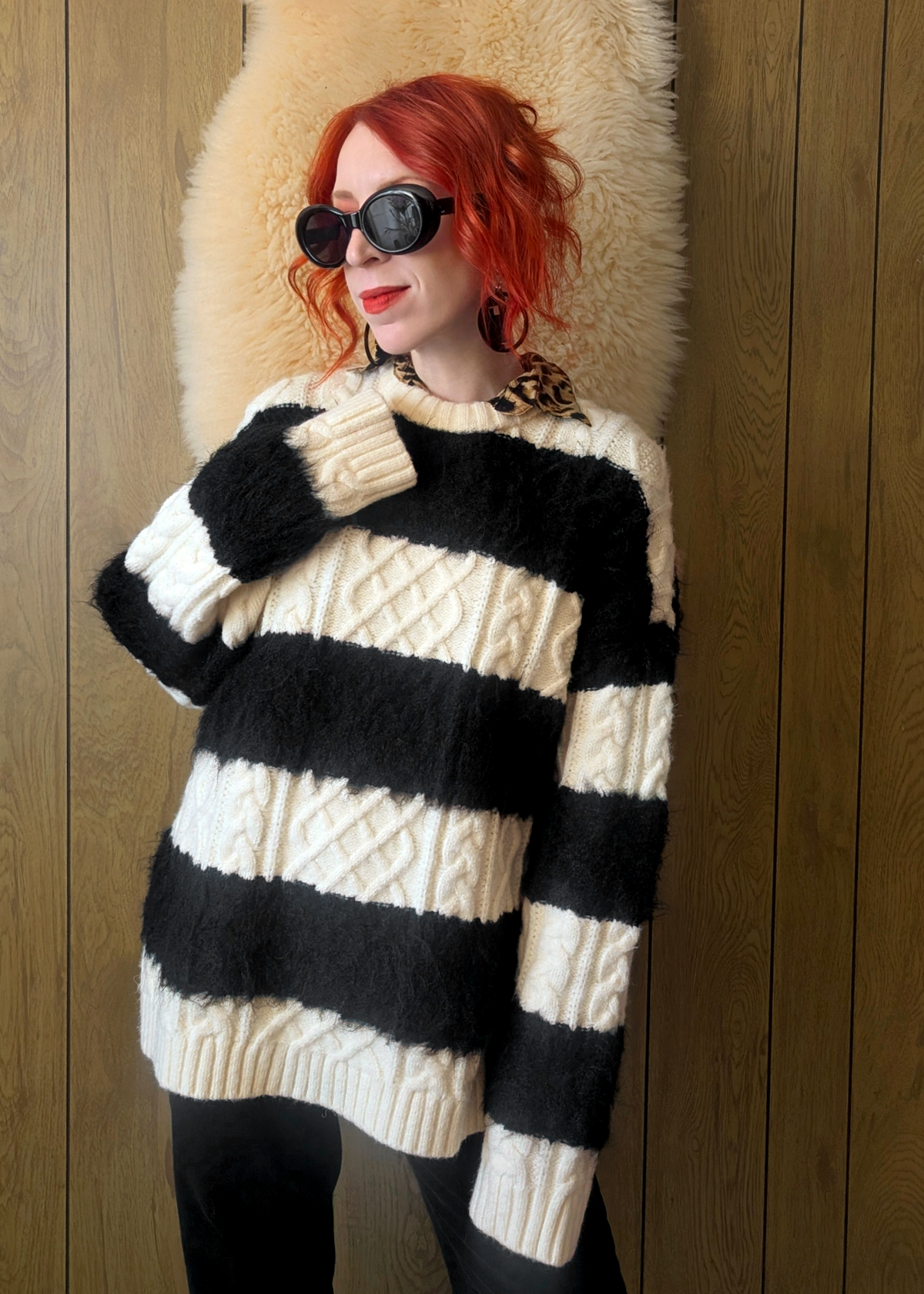 Person wearing a black and cream striped cable knit sweater layered over a leopard print collared shirt, posed against a wooden wall.
