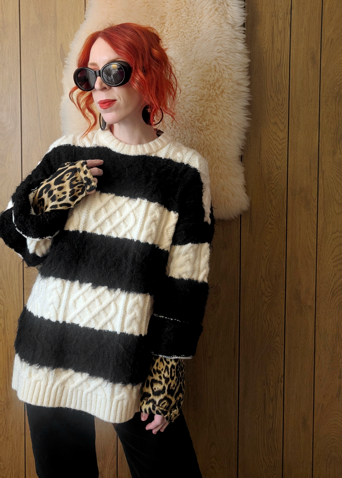Person wearing a black and cream striped cable knit sweater layered over a leopard print collared shirt, posed against a wooden wall.
