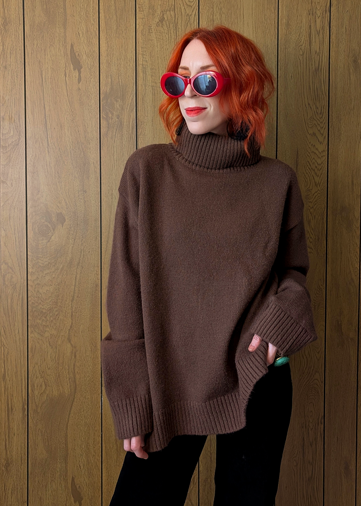 Person wearing a brown sweater and sunglasses against a wooden wall