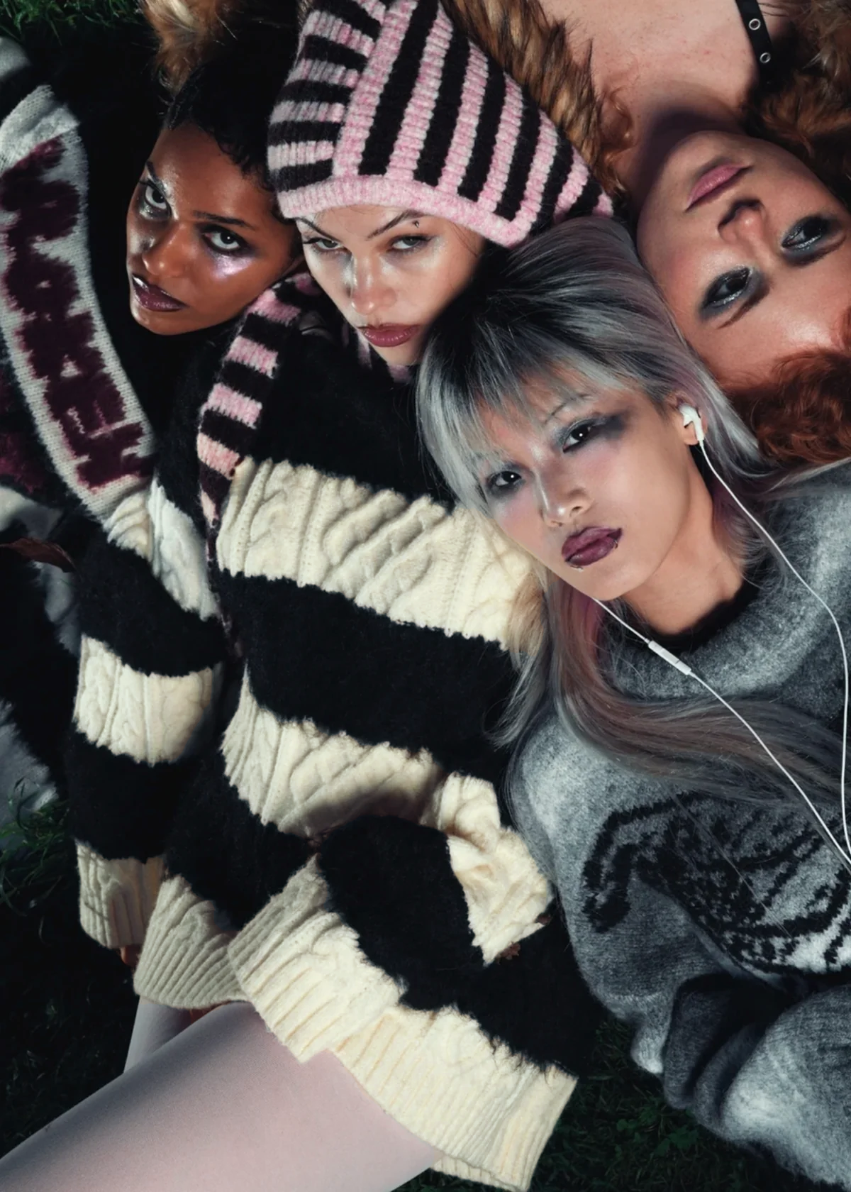 Three women wearing patterned sweaters and hats, with one listening to music on earphones. Focus is on the woman wearing a cream and black cable knit sweater as a dress with pale pink tights