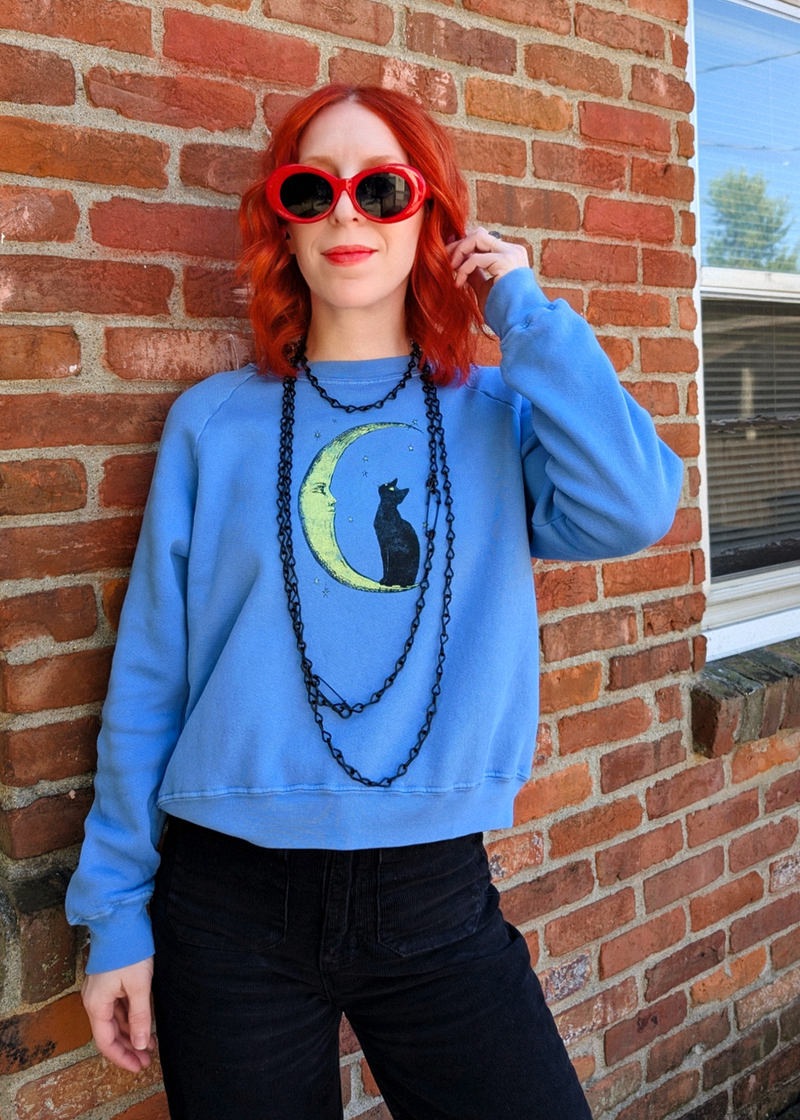 A woman with red hair wearing a light blue crewneck sweatshirt with a black cat and green crescent moon graphic, paired with black jeans, layered black chain necklaces, and red sunglasses, posing against a red brick wall.