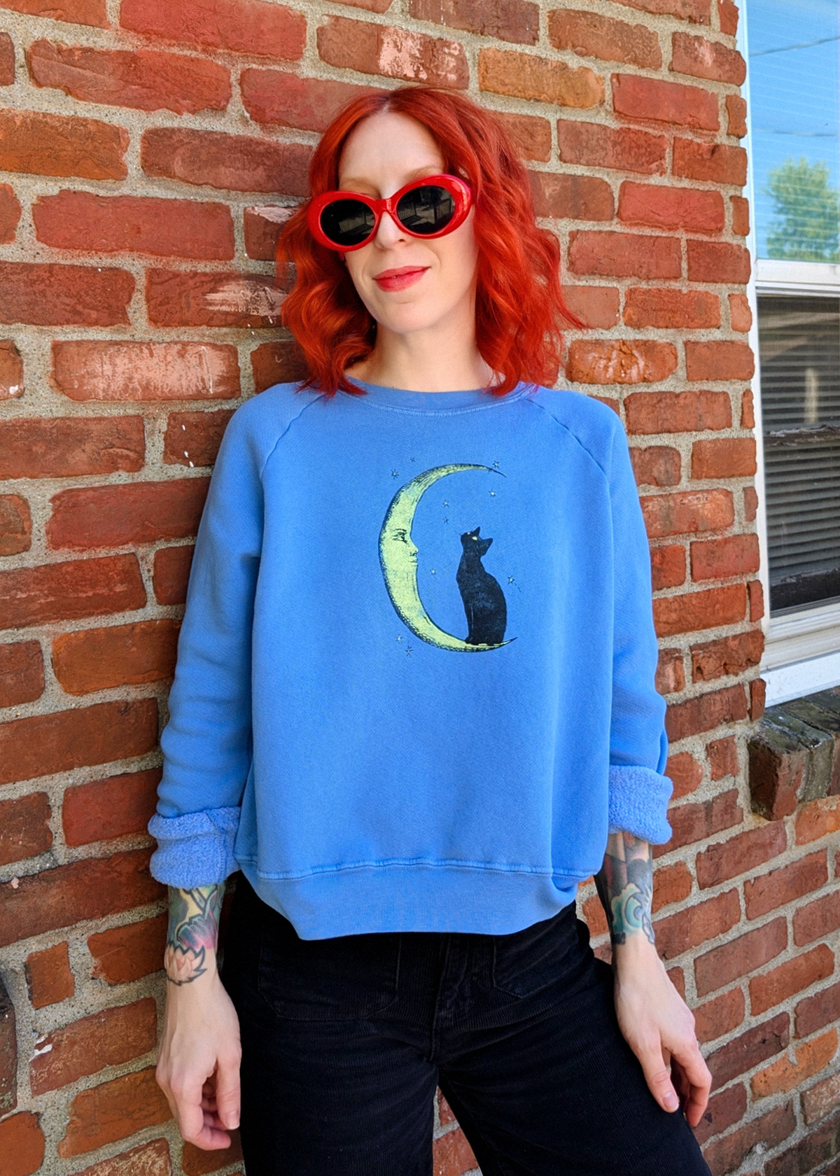 A person with red hair and red sunglasses stands against a red brick wall wearing a light blue sweatshirt with a crescent moon and cat graphic, paired with black pants.
