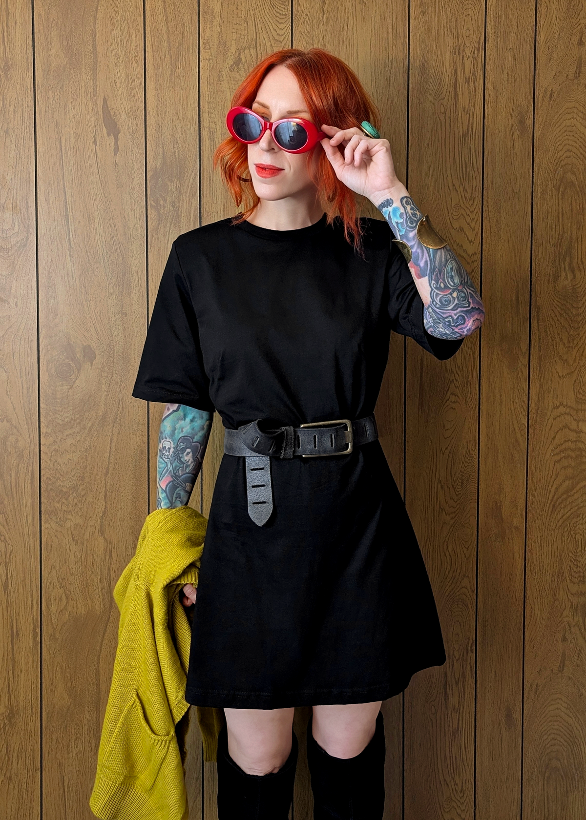 Person wearing a black dress with tattoos and colorful sunglasses against a wooden wall.