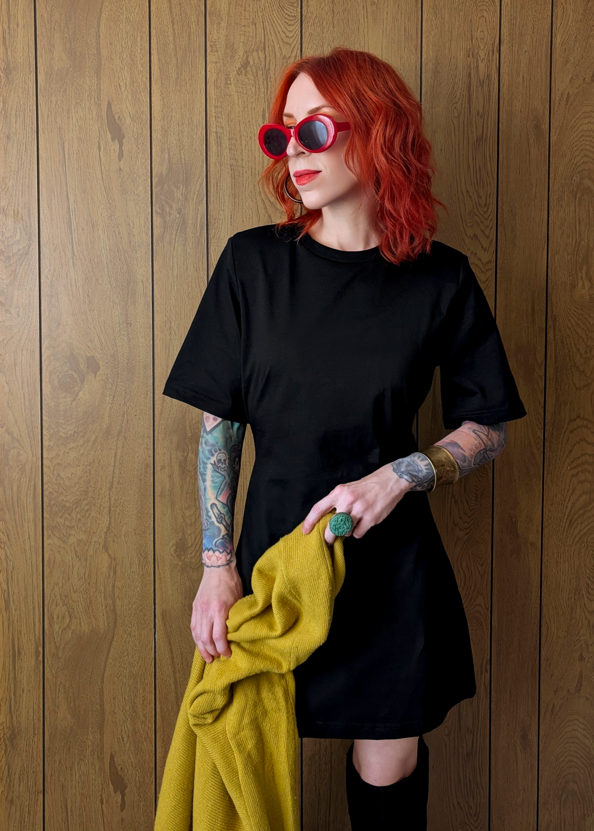 Woman with red hair wearing a black dress and sunglasses, holding a yellow jacket against a wooden wall.