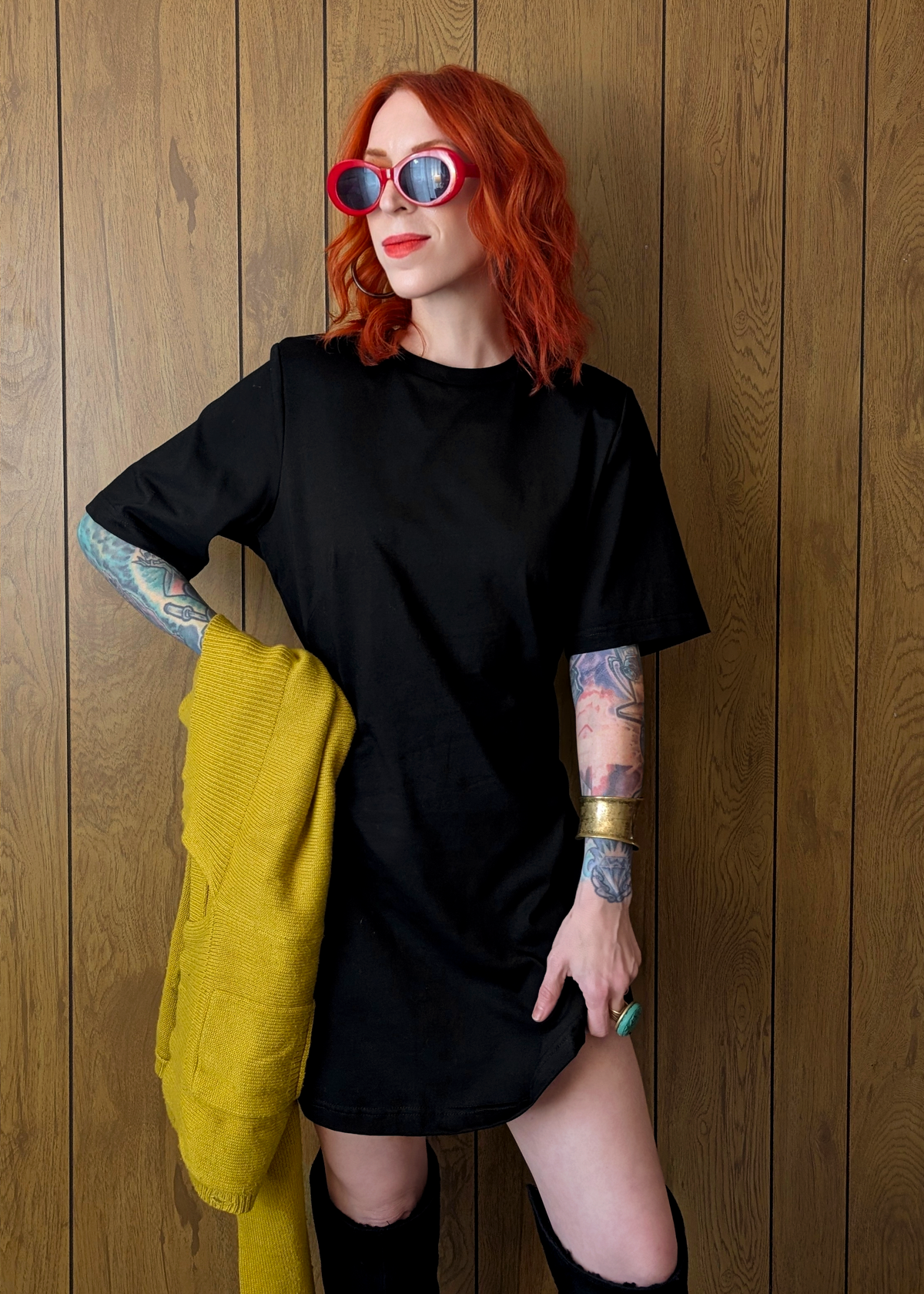 Person wearing a black outfit with tattoos and colorful sunglasses against a wooden background
