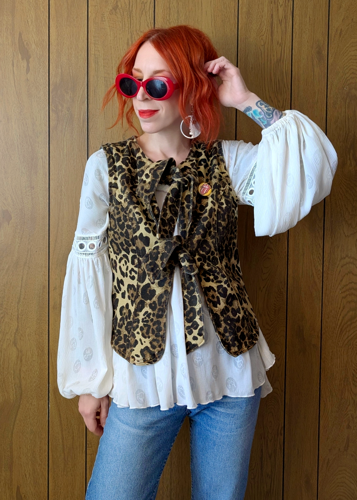 Person wearing a leopard print vest over a white blouse with blue jeans against a wooden background