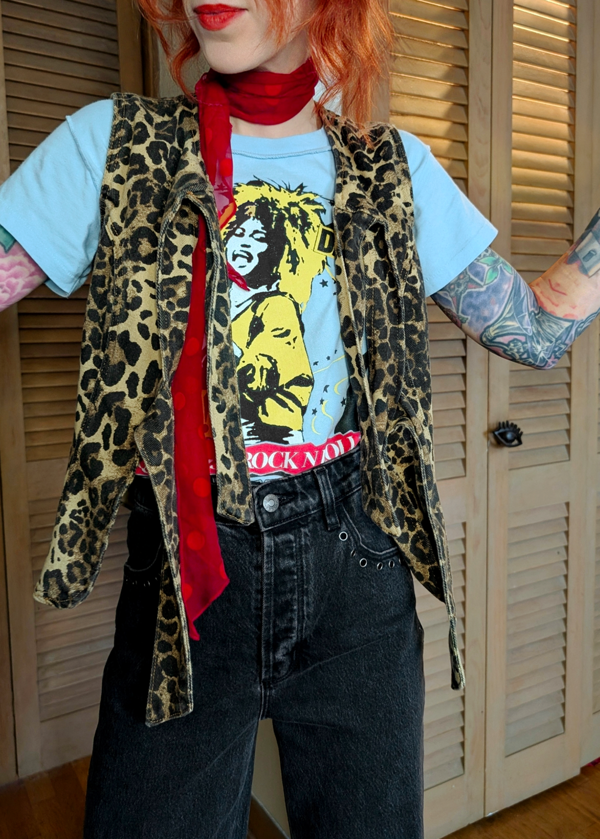 Person wearing a leopard print vest over a Tina Turner  t-shirt with a red scarf, standing in front of wooden shutters.