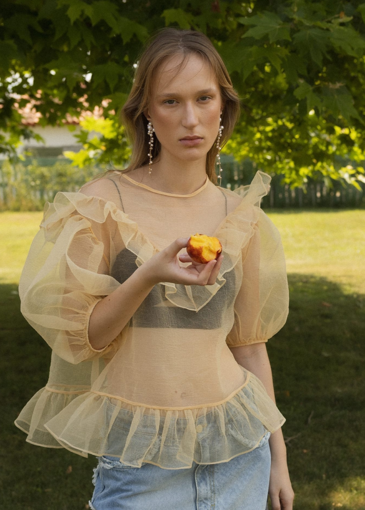 Woman wearing a sheer yellow organza balloon sleeve top over a dark bralette with denim jeans, holding an apple outdoors 