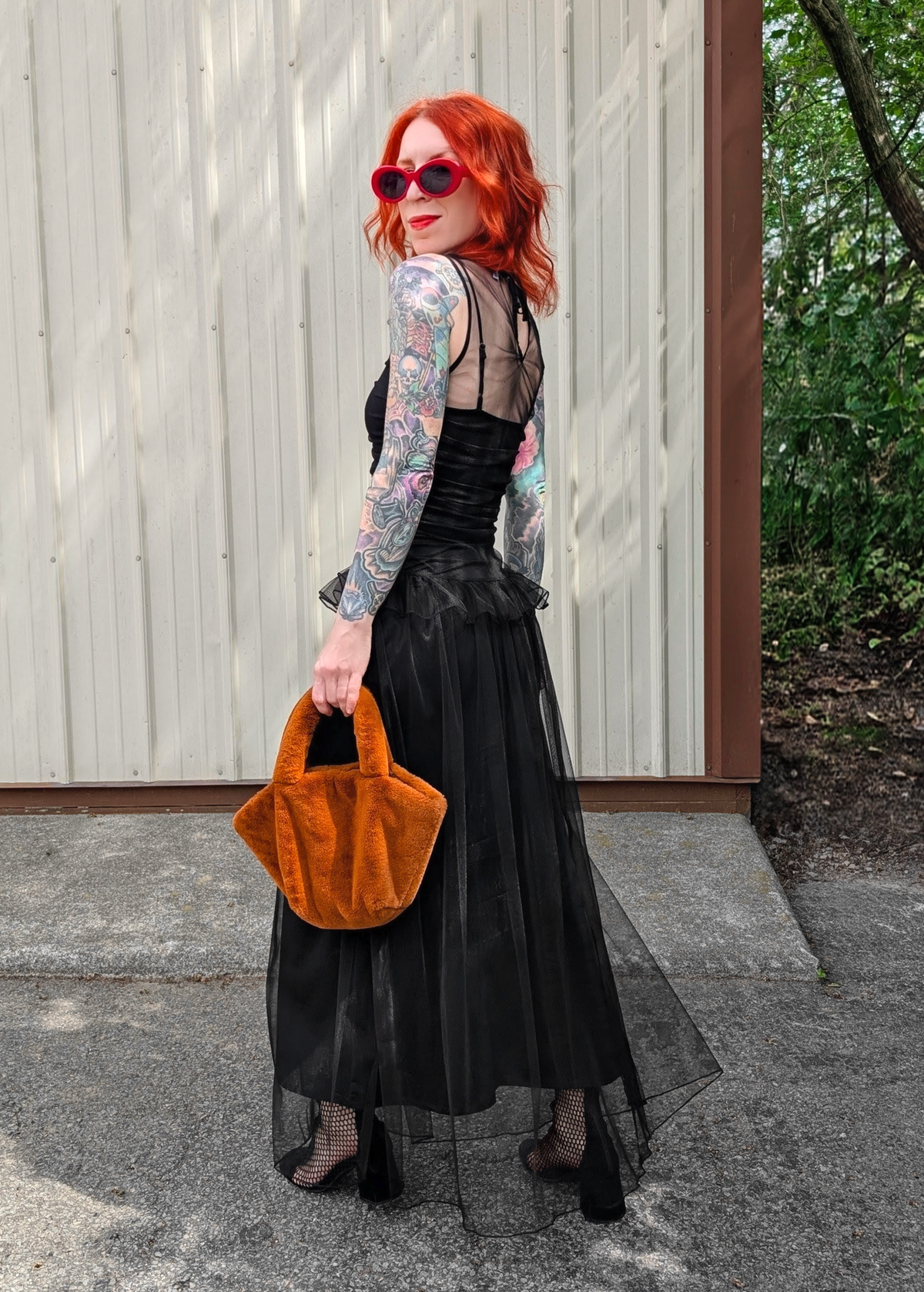 Woman with red hair wearing a black  tulle dress holding a rust brown faux fur handbag outside