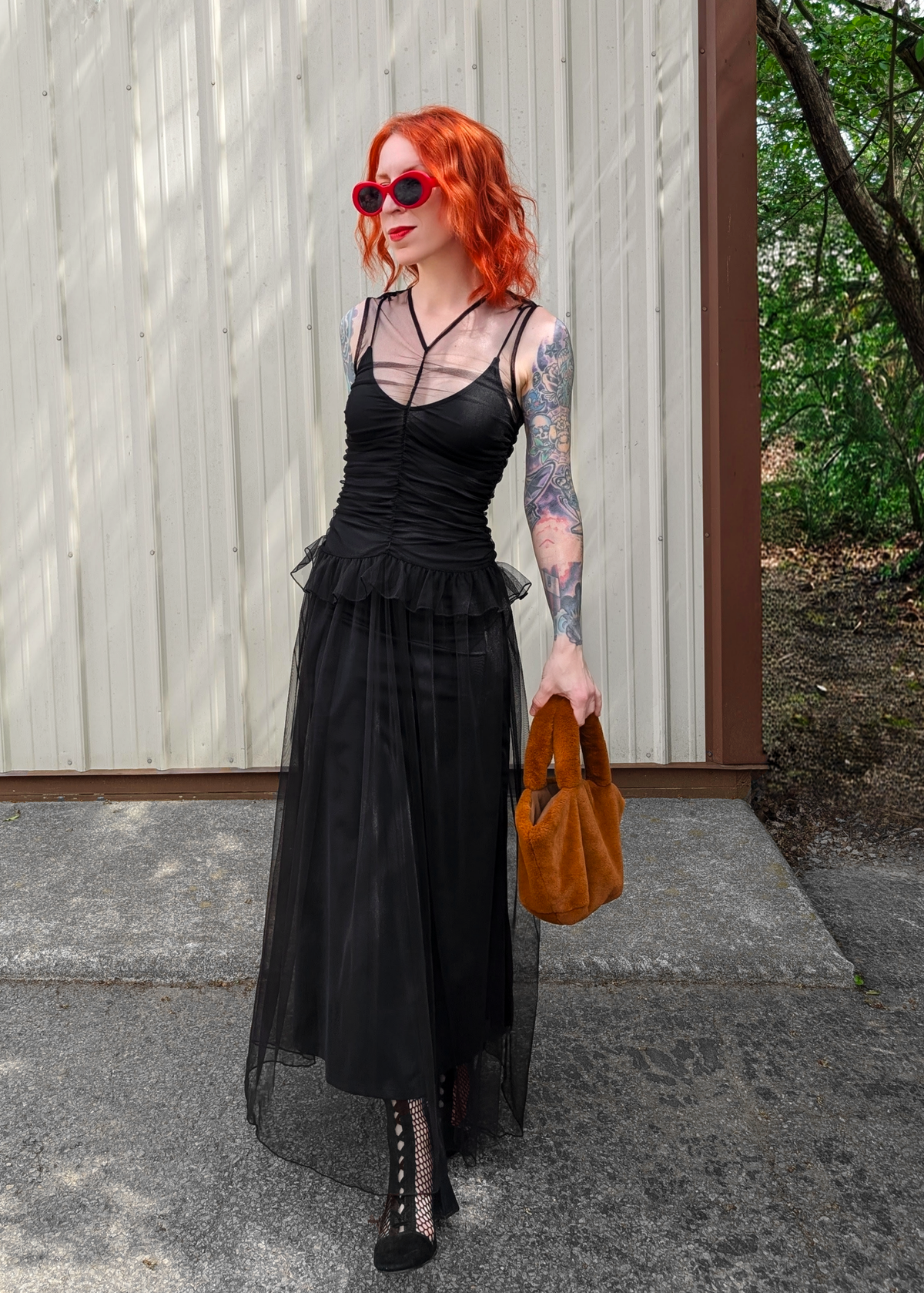 Person wearing a black tulle dress with sheer elements, holding a brown faux fur handbag, standing outdoors.