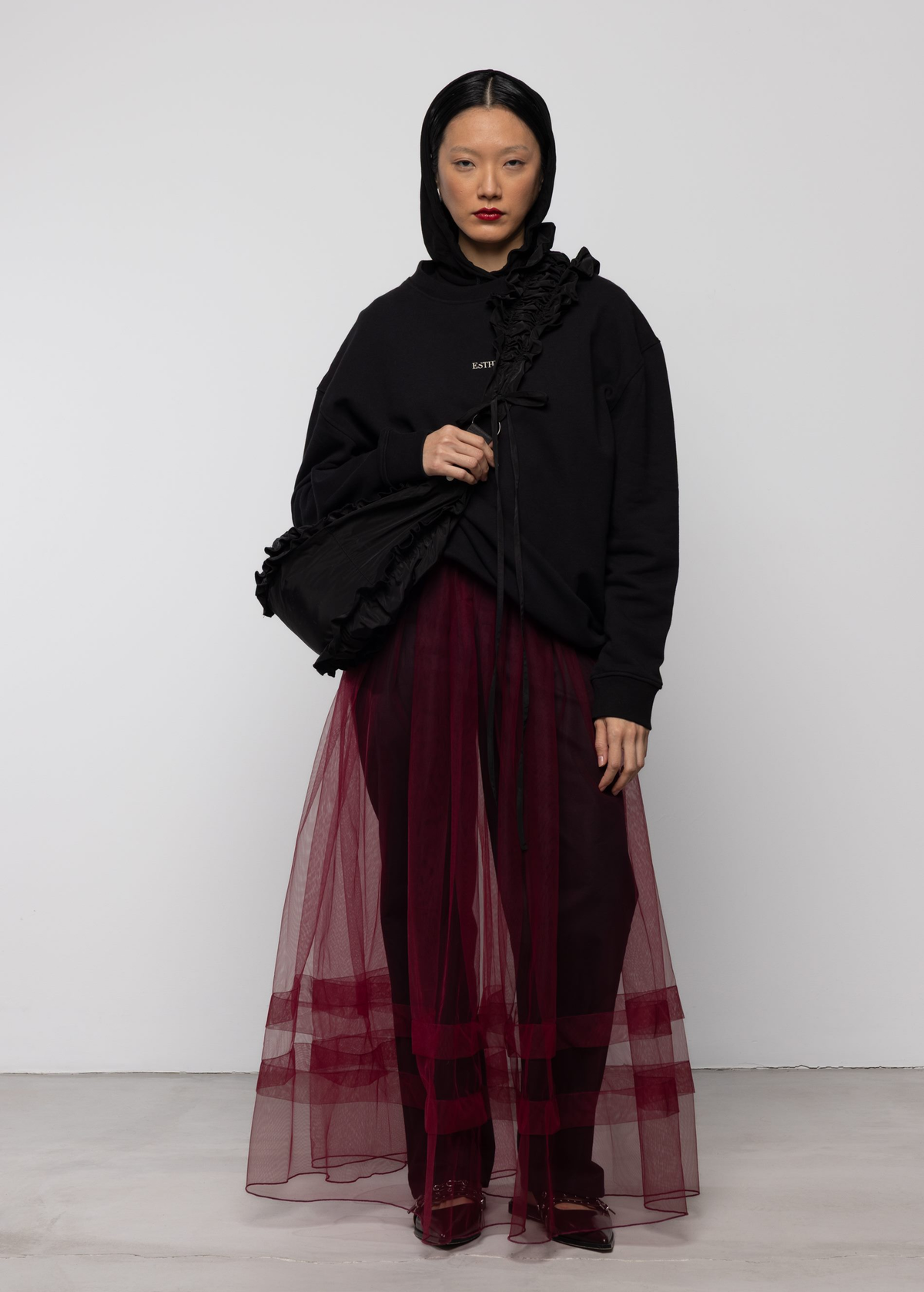 Woman wearing a sheer burgundy tulle dress and black hoodie and black trousers against a white background