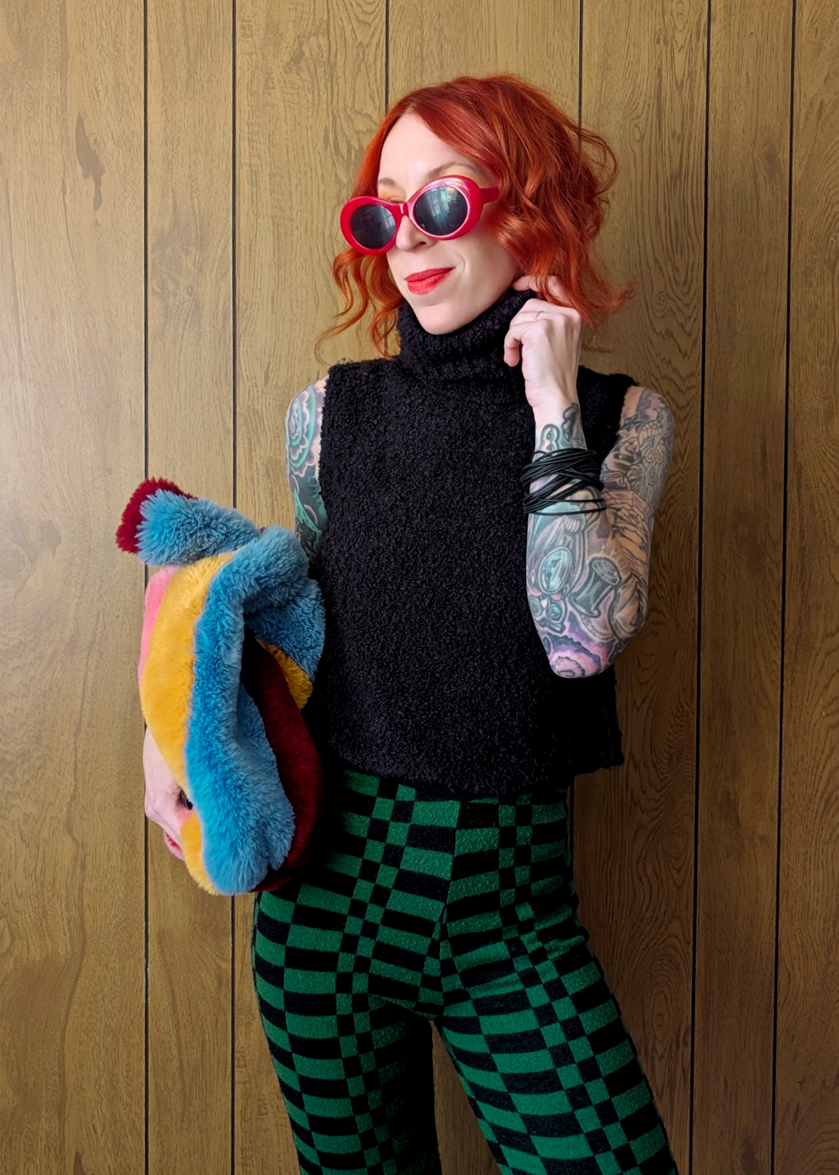 Person with red hair wearing sunglasses, a black sleeveless knit top, and checkered pants against a wooden wall.
