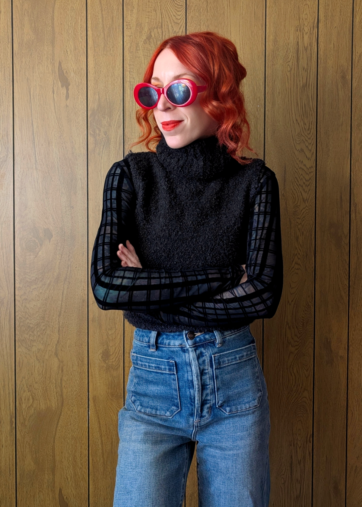 Person wearing a black turtleneck vest over a sheer sleeved top and blue jeans against a wooden paneled wall.