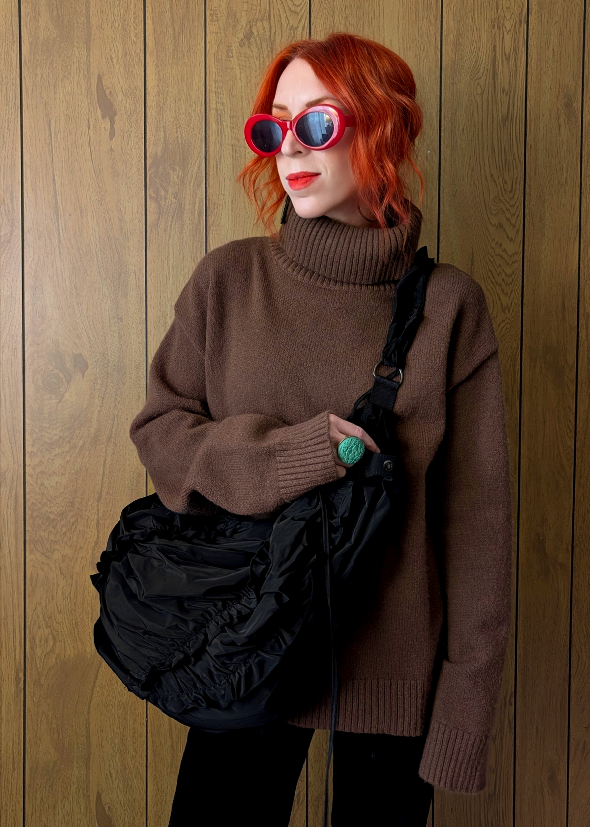 Person wearing a brown sweater and sunglasses, holding a black bag against a wooden background