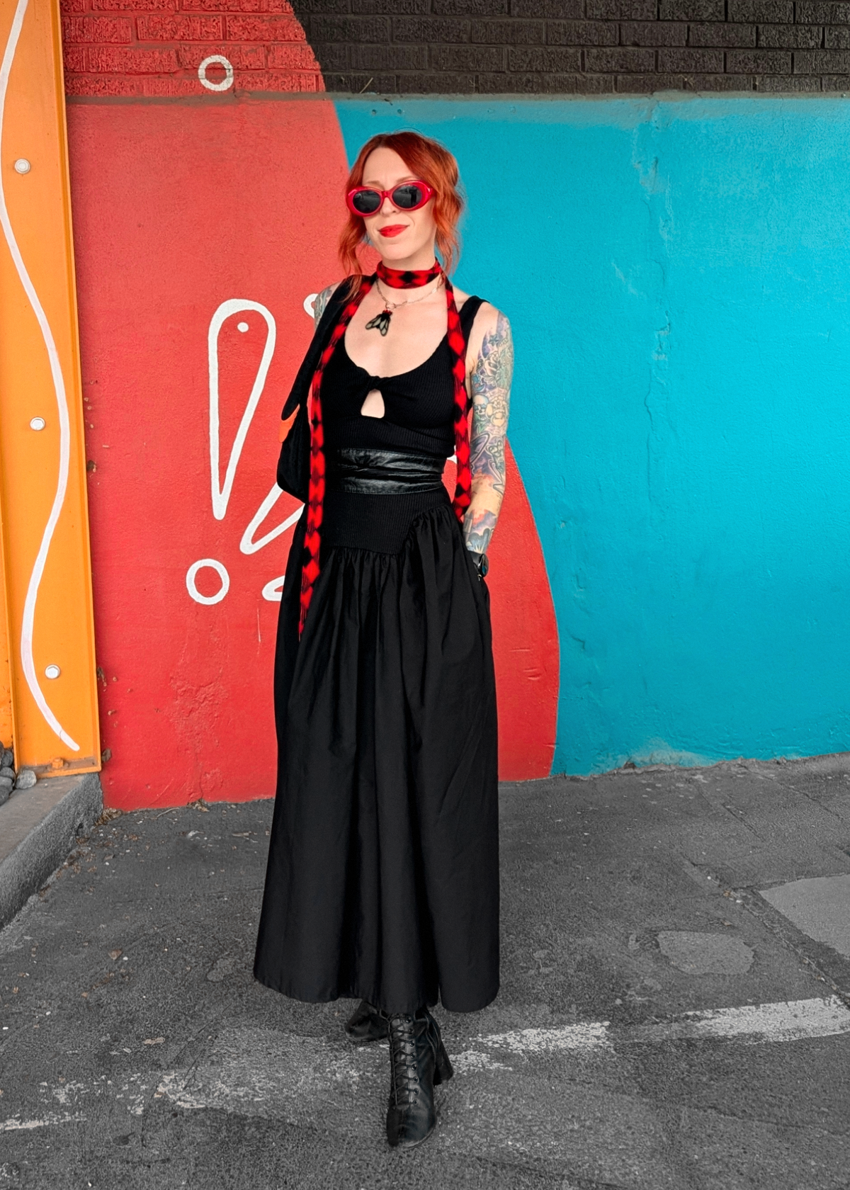 Person wearing a black dress, belted at waist and wearing a red scarf standing against a colorful wall 