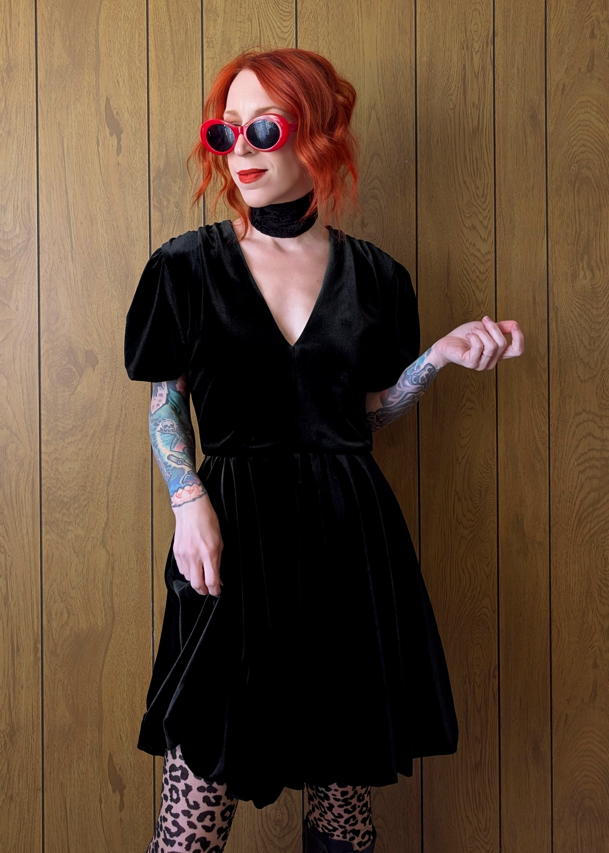 Person wearing a black velvet babydoll dress with red sunglasses against a wooden panel background
