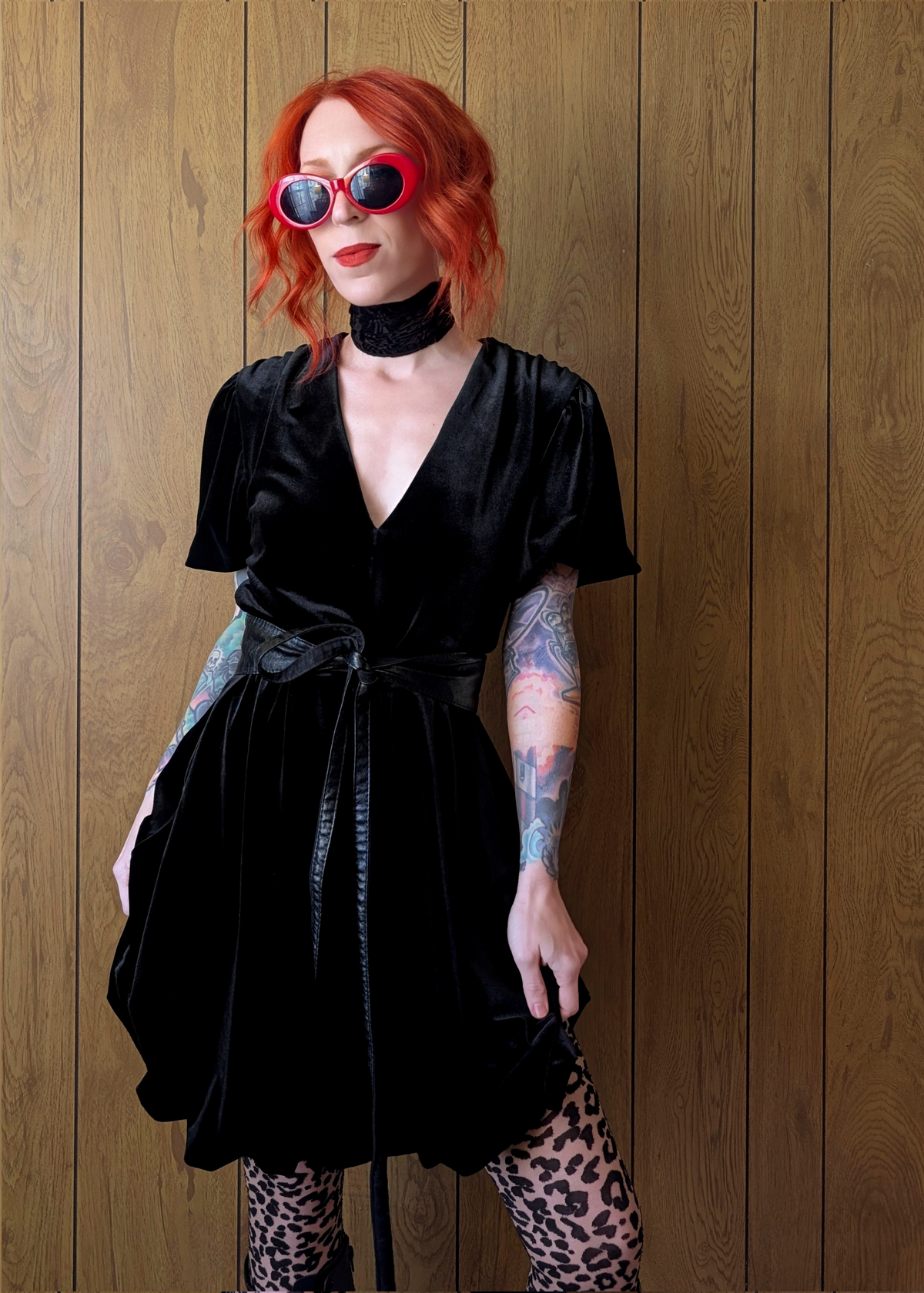 Person wearing a black velvet mini dress with leopard print tights against a wooden panel background. Dress is belted at the waist with a black tie belt