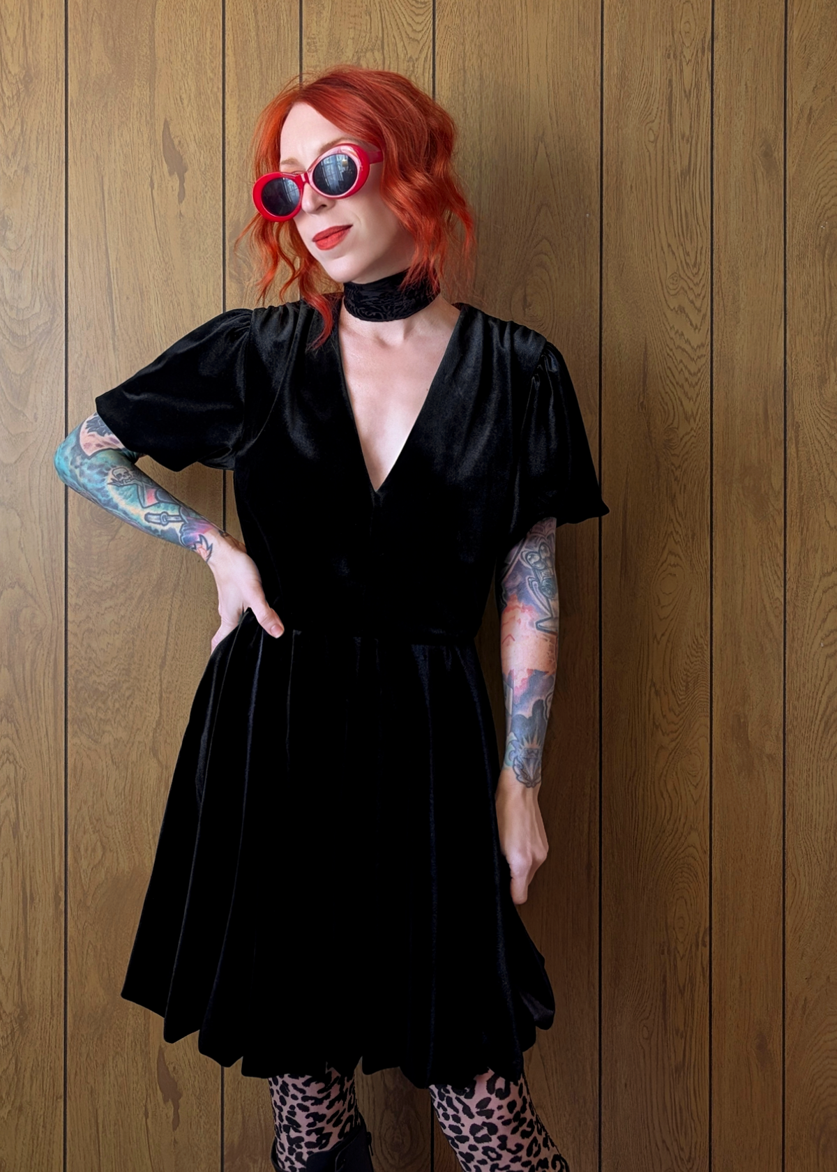 Person wearing a black velvet babydoll dress with red hair and tattoos standing against a wooden wall.