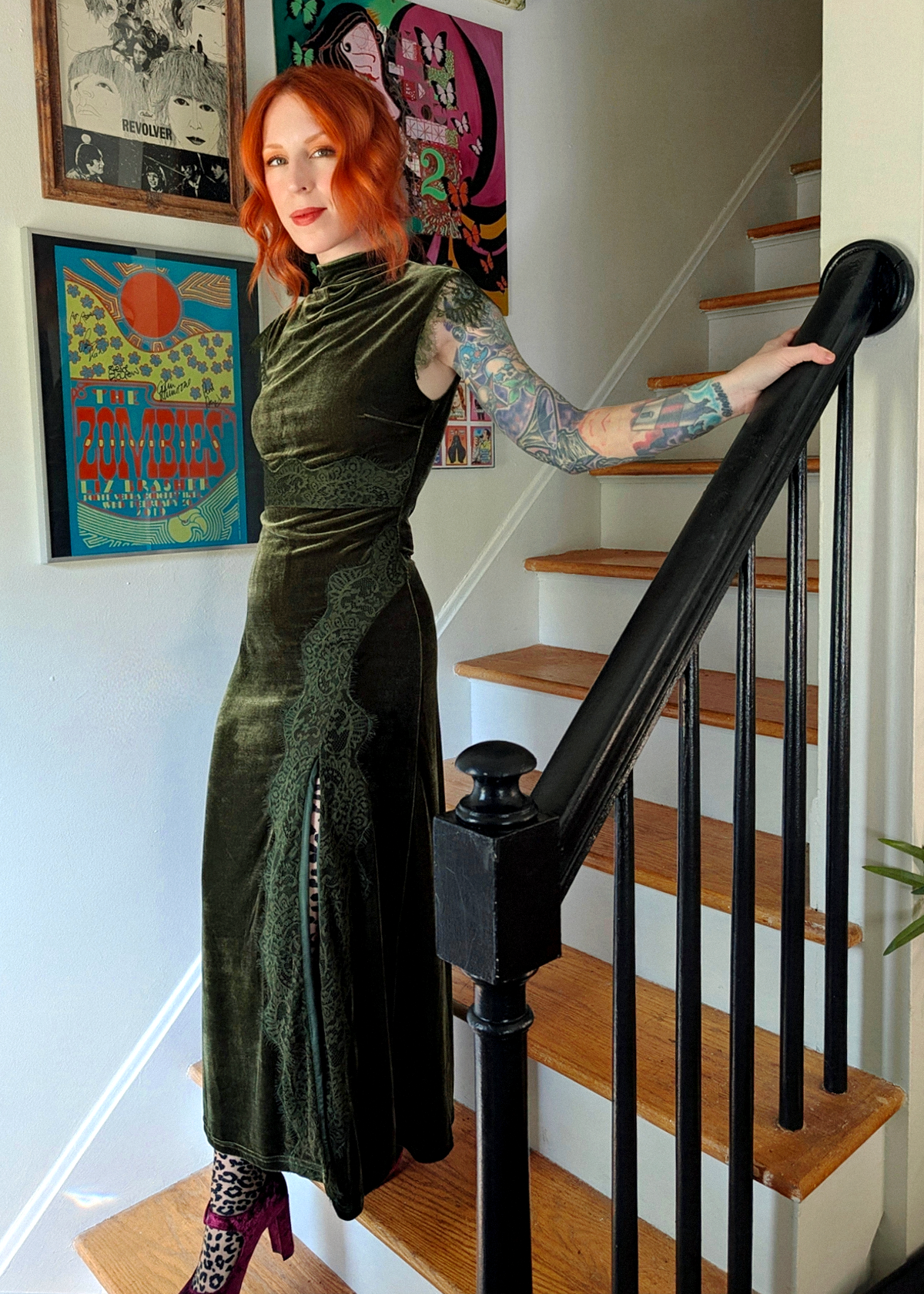 A woman with red hair stands on wooden stairs holding a black handrail, wearing a dark olive green velvet sleeveless dress with lace detailing, styled with leopard print tights and burgundy velvet heels. Framed posters hang on the wall behind her.