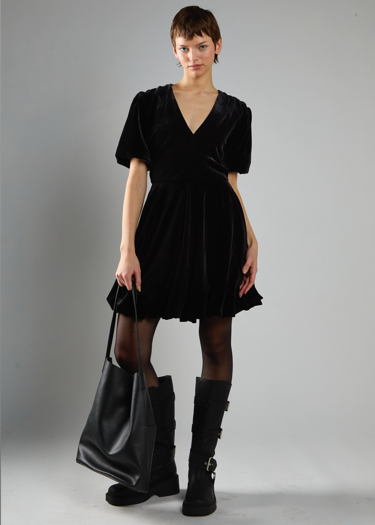 Woman wearing a black velvet mini dress with short sleeves and a V-neckline, holding a black handbag on a gray background.
